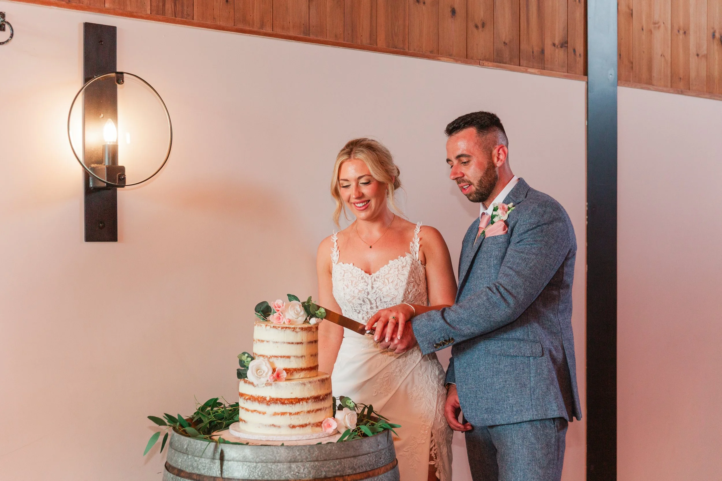 A bride in a lace dress cutting their 2 tier naked wedding cake with her groom dressed in a blue suit