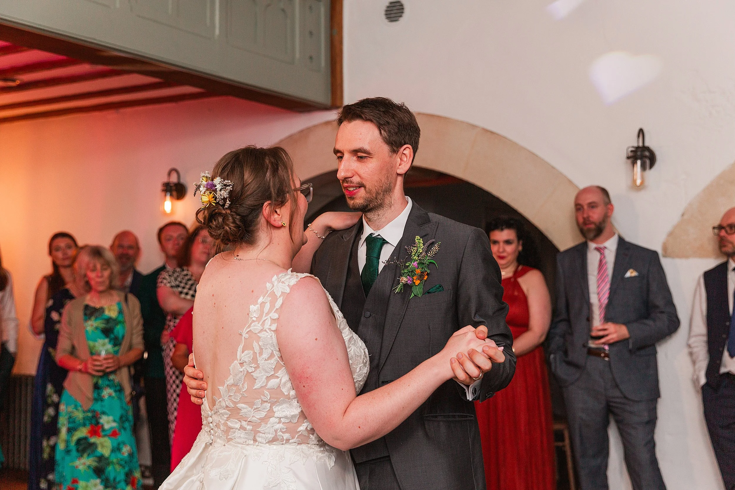 A groom with dark hair wering a grey suit and green tie dancing with his bride in a barn wedding venue in front of their wedding guests
