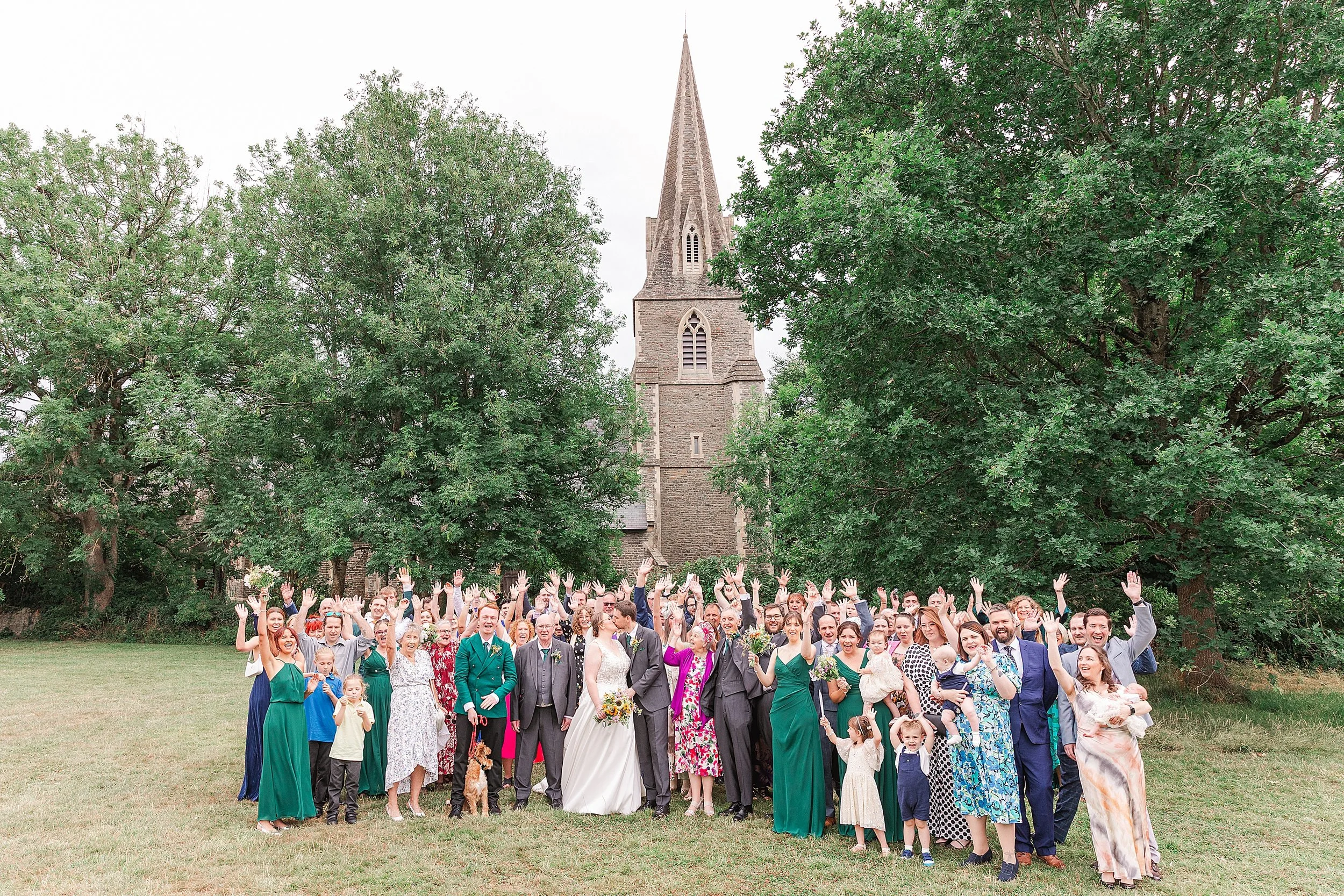 A bride with brown hair and glasses standing with her groom in a grey suit with all their wedding guests cheering outside their church