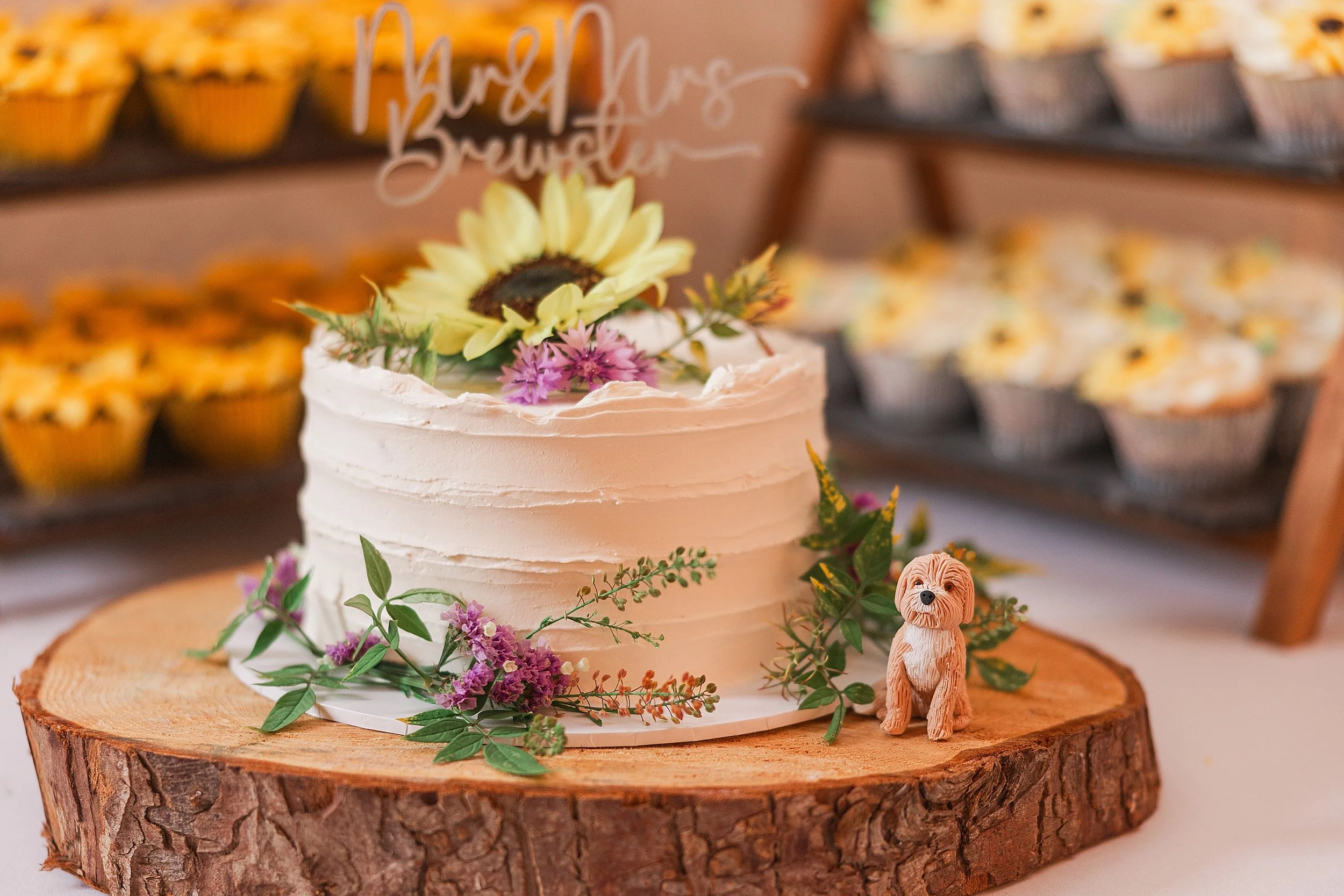 A one tier white wedding cake on a wooden log slice with a sunflower on top and a little god character sitting on the edge