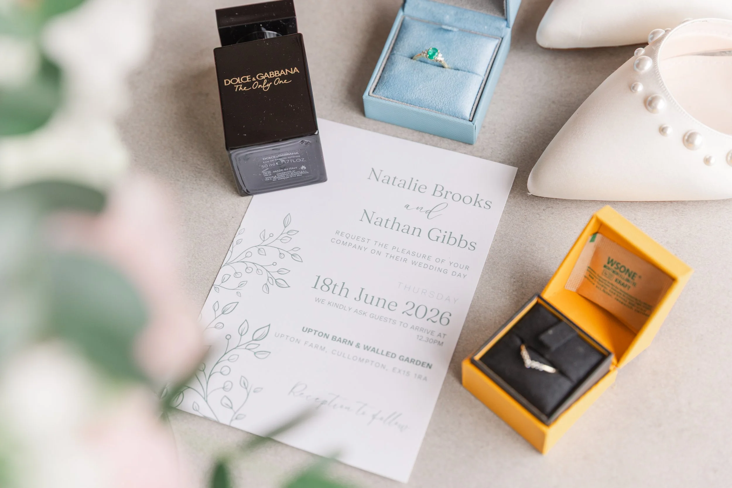 A pretty flat lay of a wedding invite with wedding details laid next to it including an engagement ring and a bottle of dolce and gabbana perfume