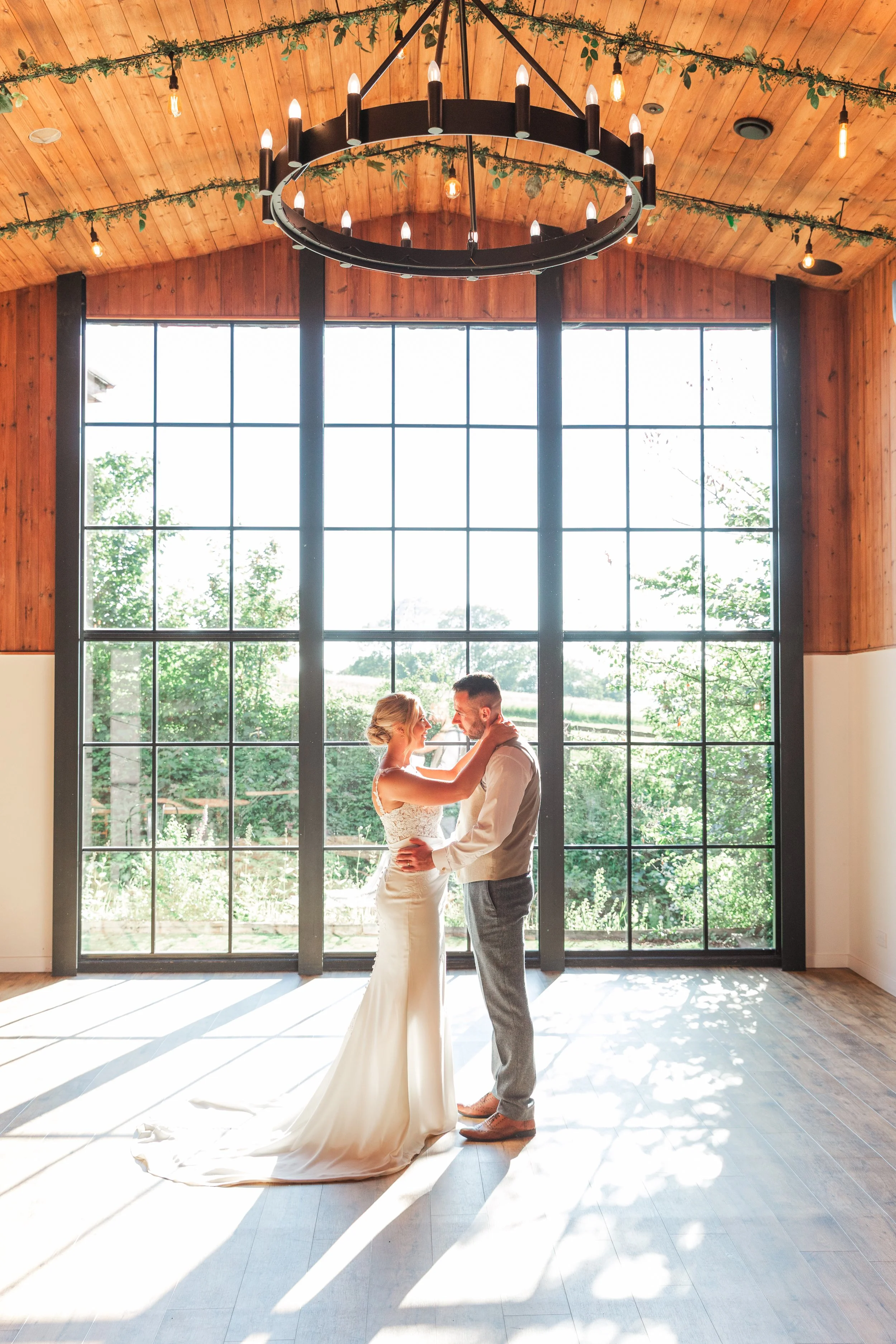 A bride in a lace dress standing facing her groom in a blue suit in front of a floor to ceiling window underneath a black chandelier with candlelights