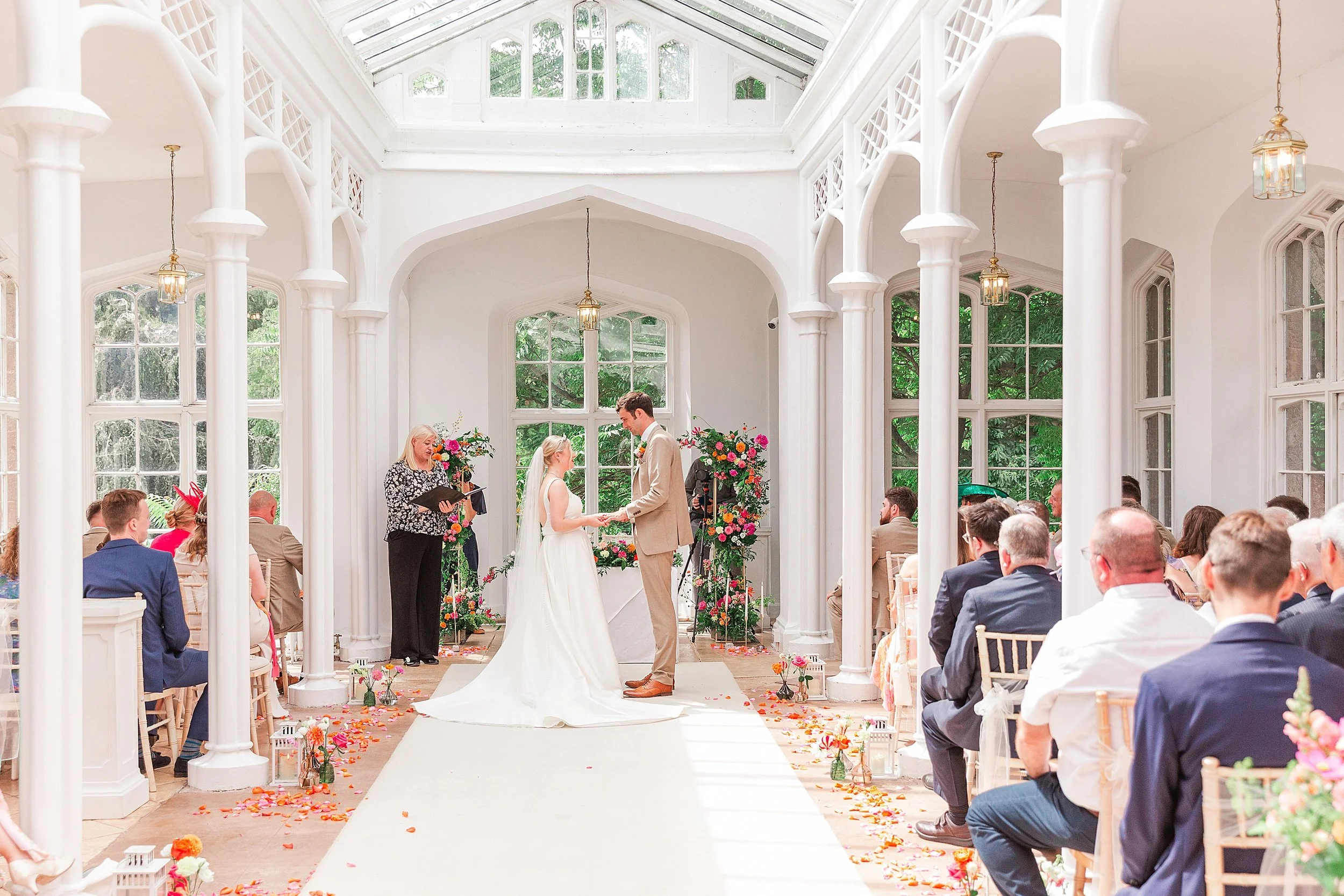 A bride and groom in a white orangery saying their vows surrounded by colourful florals