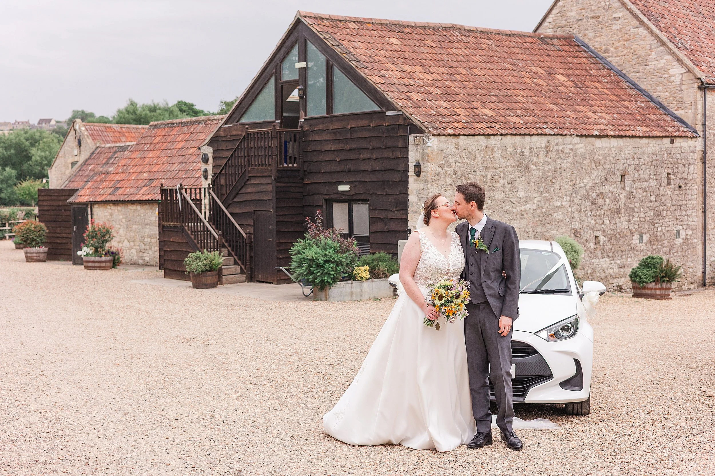A bride with brown hair and glasses kissing her groom in a grey suit standing in front os a white car with a stone farmhouse building behind them
