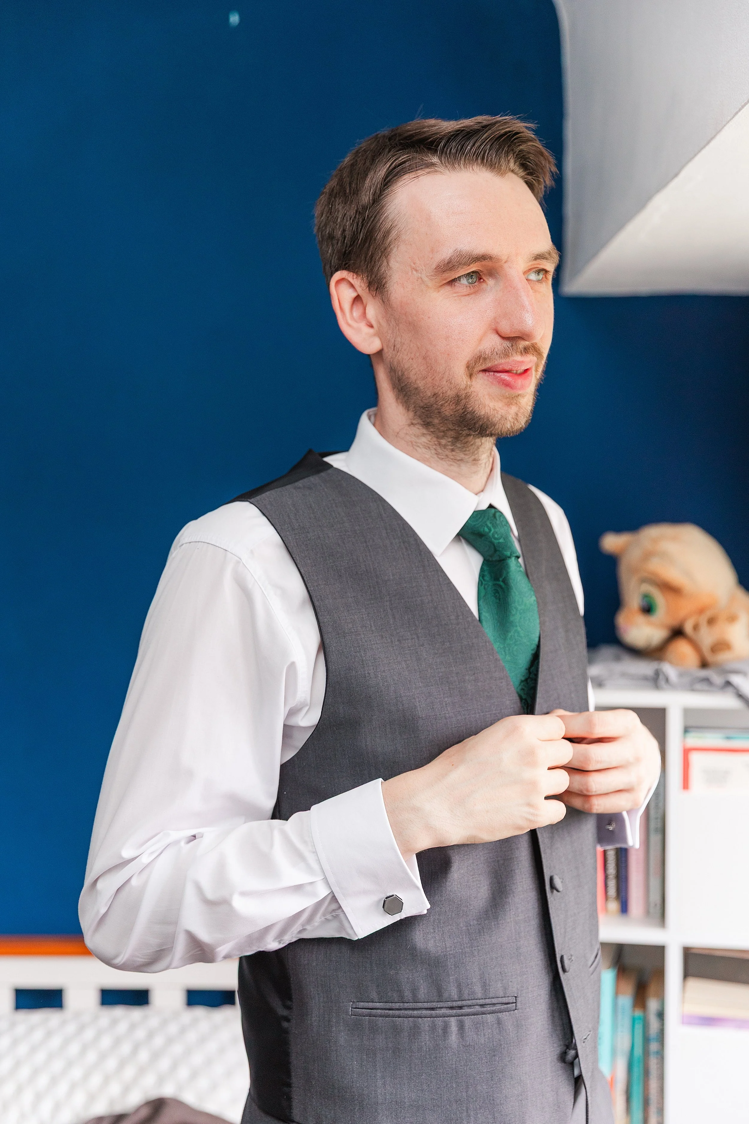 A groom standing in a room with blue walls tying his green tie looking out hte window, he is wearing a grey waistcoat