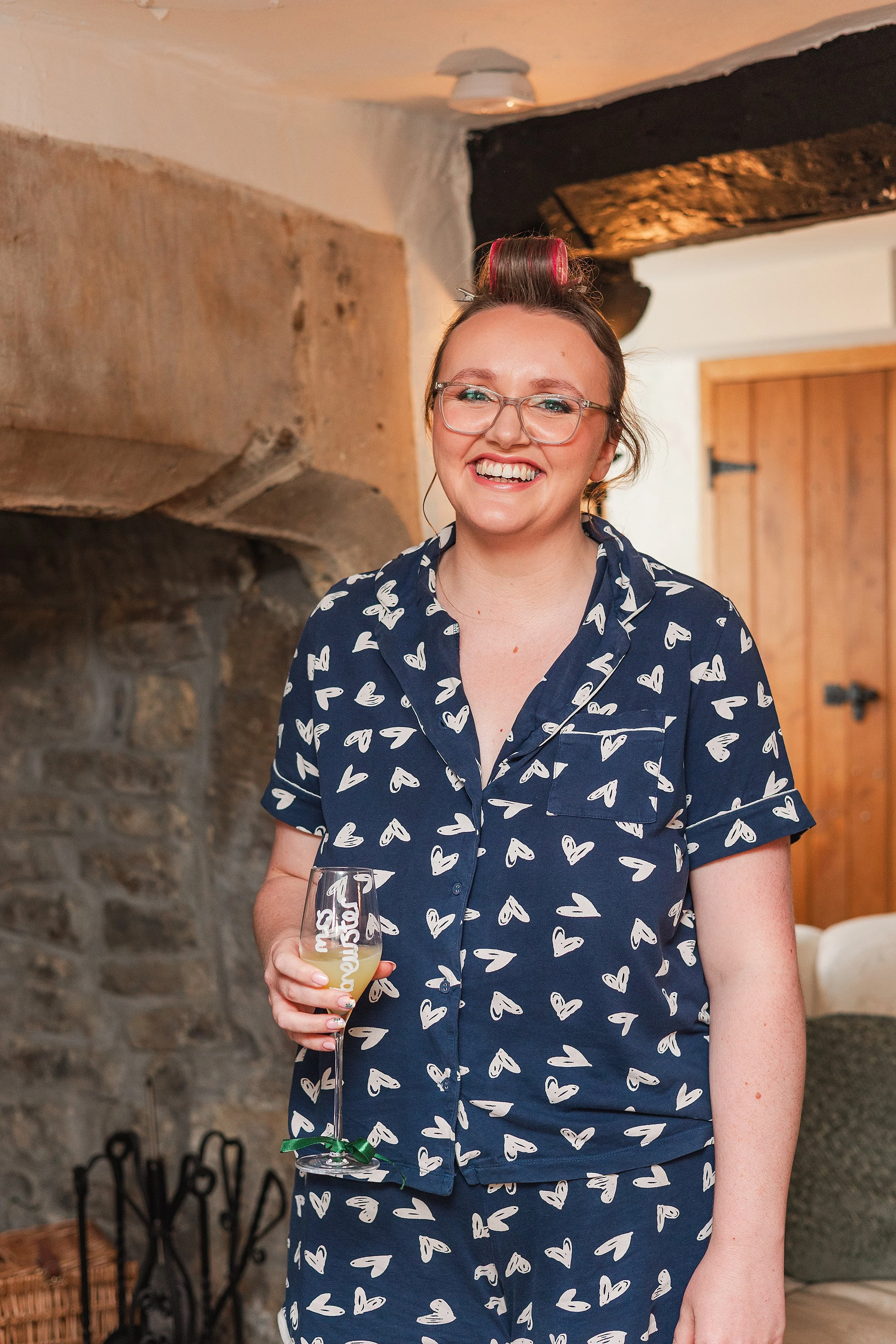 A bride with brown hair and glasses smiling at the camera, she is dressed in navy pyjamas with white hearts and holding a glass of orange juice