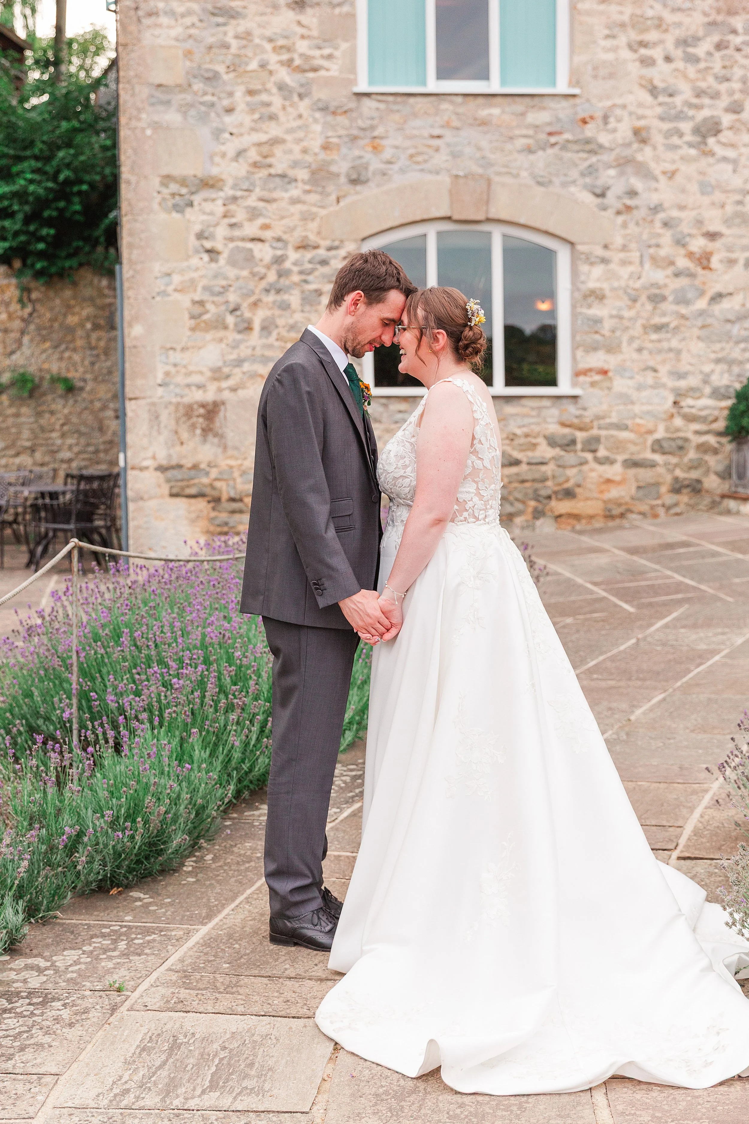 A bride with brown hair and glasses leaning her forehead againgst her groom with dark hair and a grey suit in front of a stone brick wedding venue with lavender filled gardens