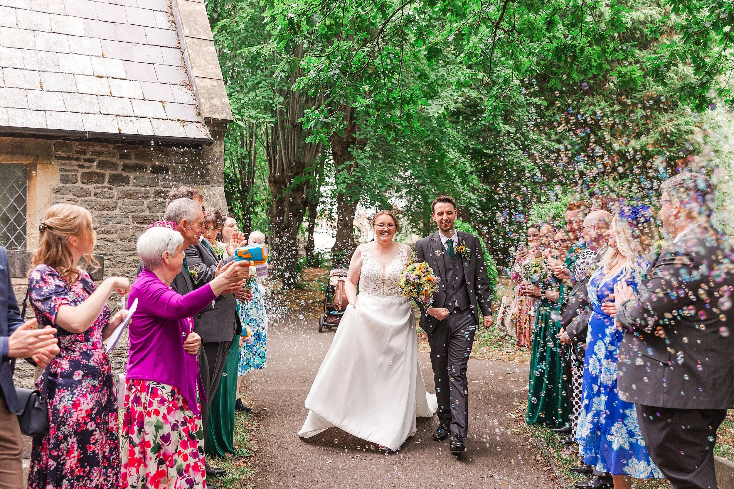 A bride with brown hair and glassed walking through abubbel confetti exit with her groom dressed in a grey suit
