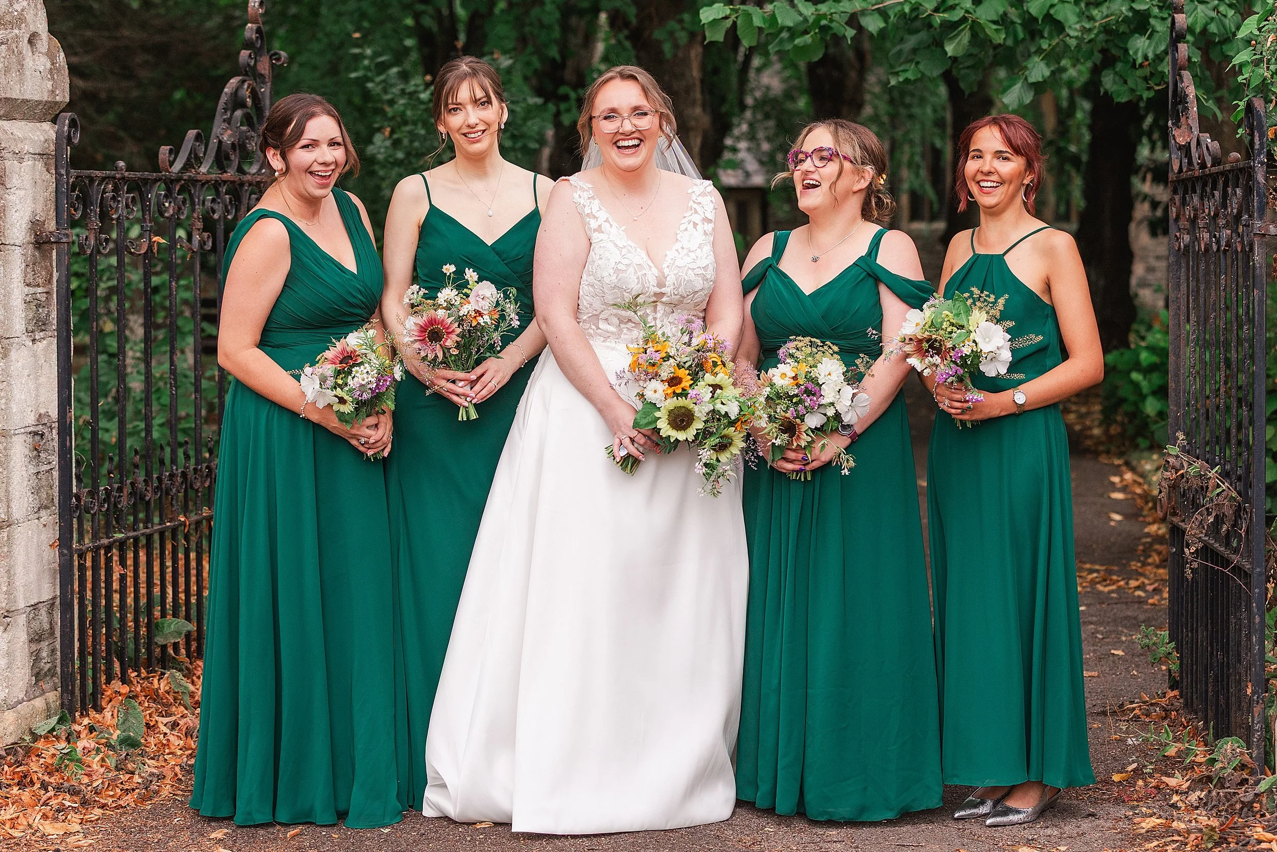 A bride with brown hair and glasses laughing with her 4 bridesmaids dressed in forrest green dresses at the gates to a church