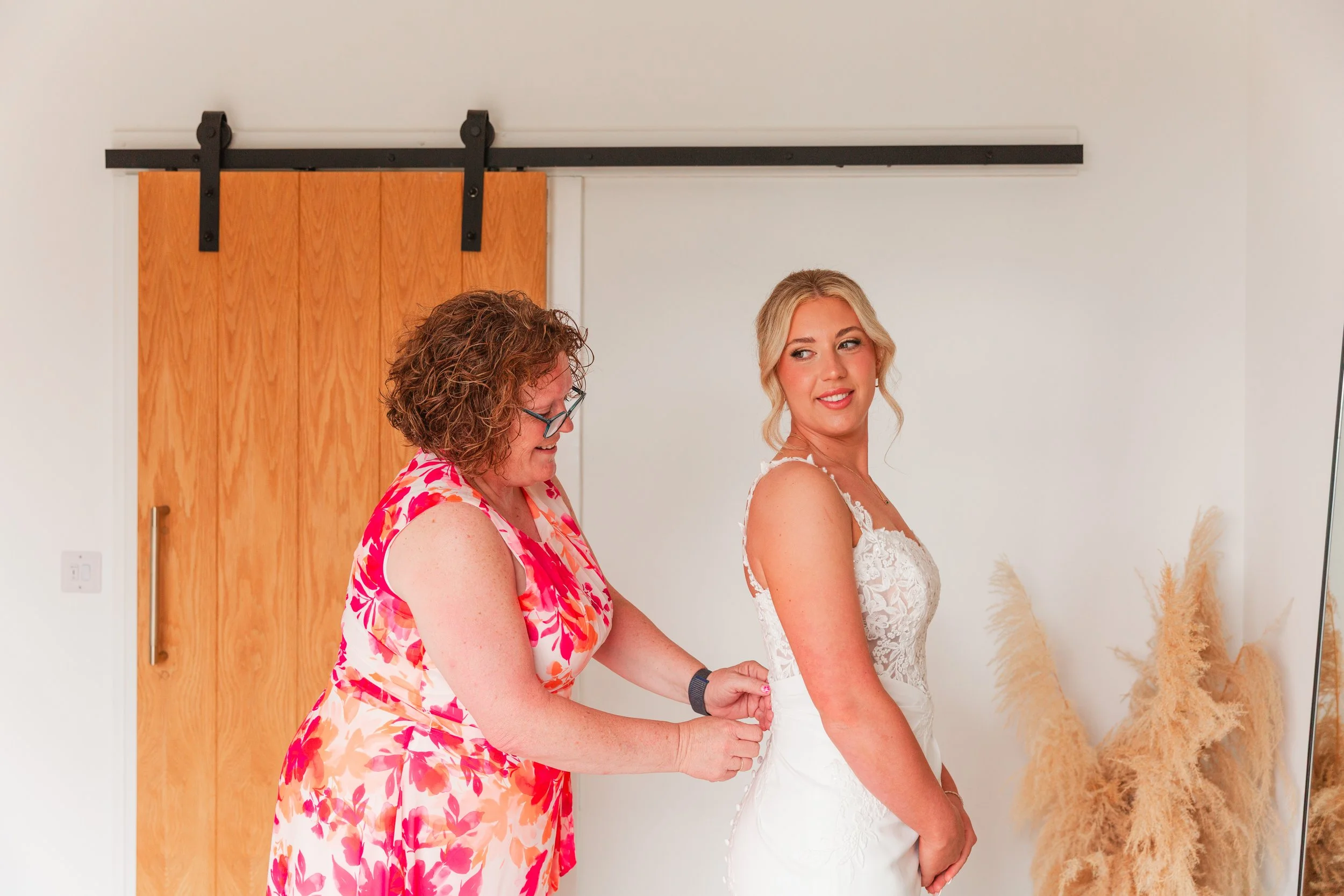 A bride having her lace wedding dress buttoned by her mum wearing a pink and white dress