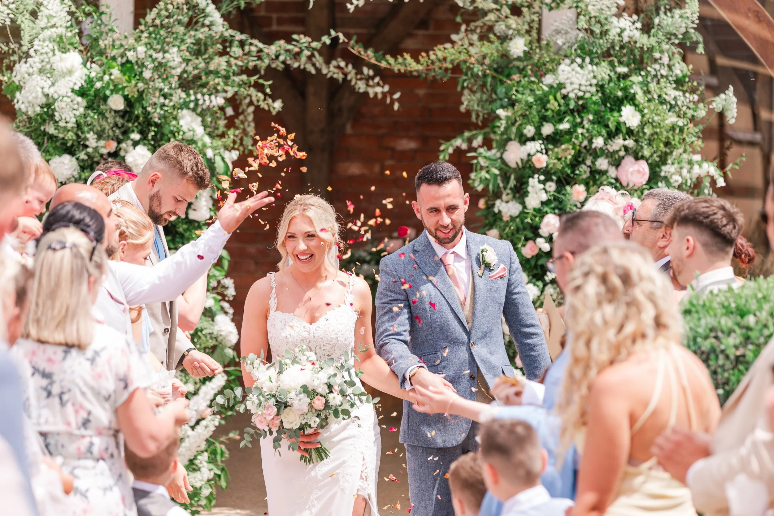 A bride in a lace dress walking through a confetti exit with her groom in a blue suit