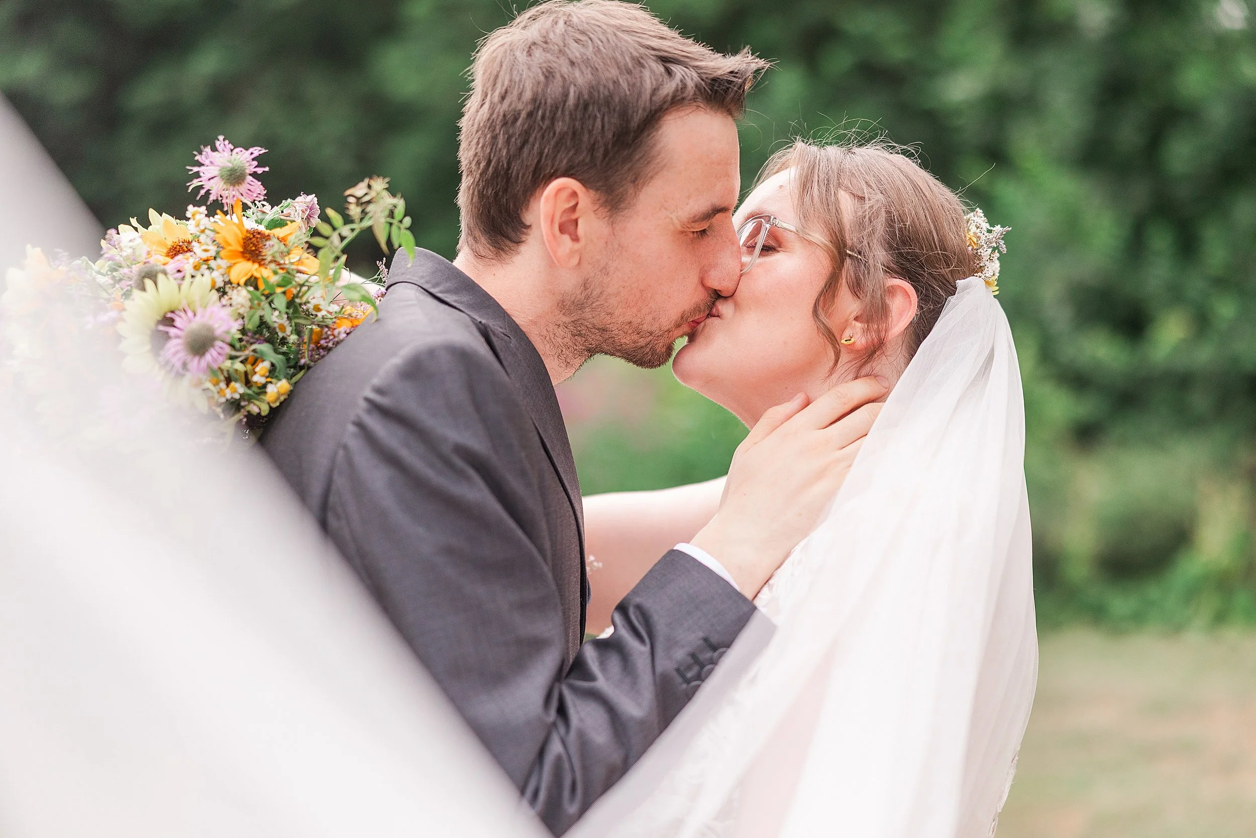 A bride with brown hair and glasses kissing her groom with dark hair and a grey suit with the wedding veil covering them