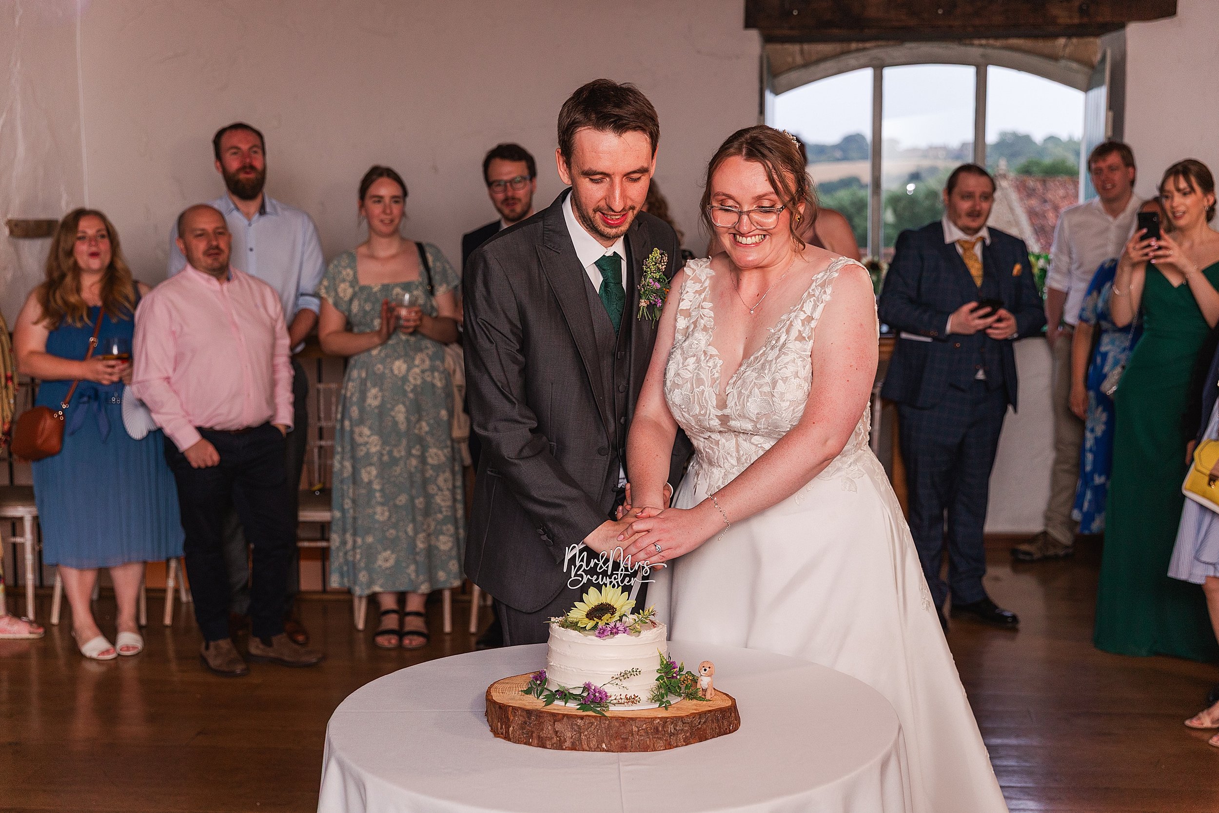 A bride with brown hair and glasses cutting her wedding cake with her groom with dark hair and a grey suit in a barn wedding venue surrounded by their wedding guests