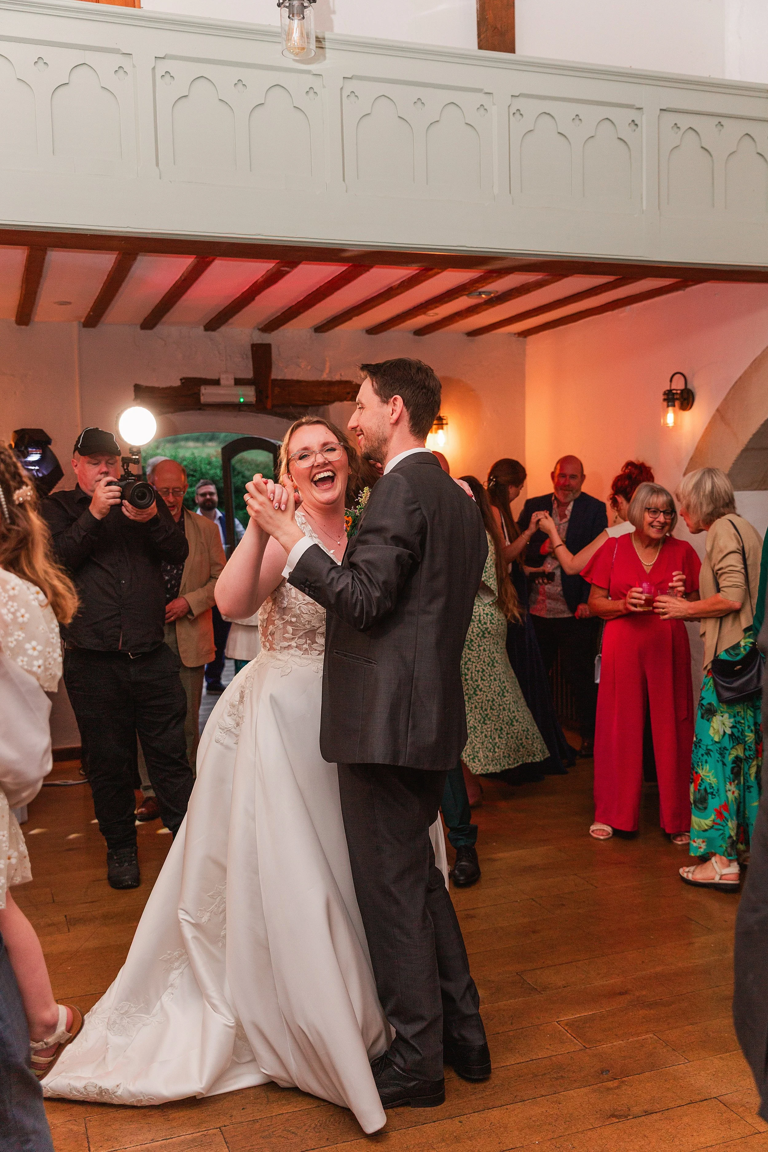 A bride with brown hair and glasses dancing with her groom with dark hair and a grey suit in a barn wedding venue