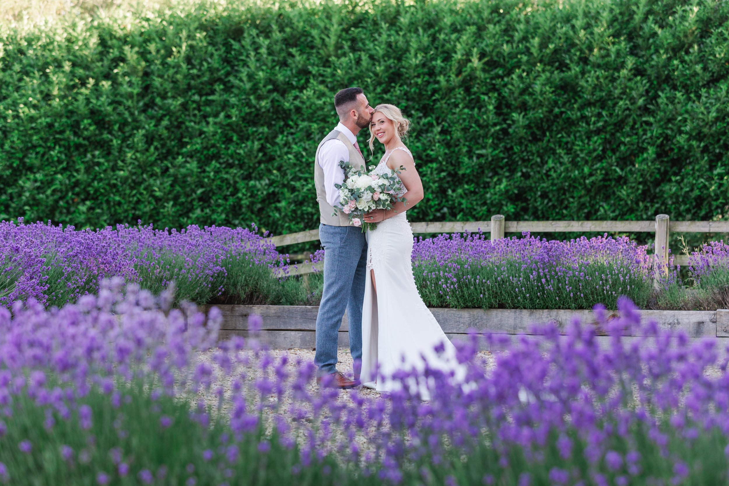 A bride in a futted lace dress standing in front of pretty lavender with her groom in a stone waistcoat kissing her forehead.