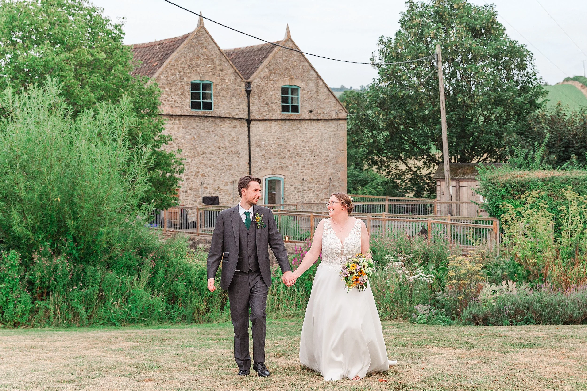 A bride with brown hair holding hands with her groom in a grey suit walking across a filed in front of a stone barn wedding venue