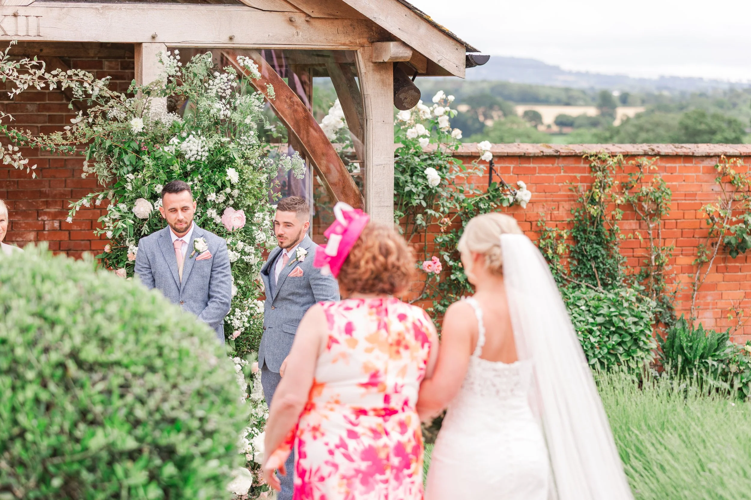 A groom in a blue suit watching his bride walk towards him down the aisle