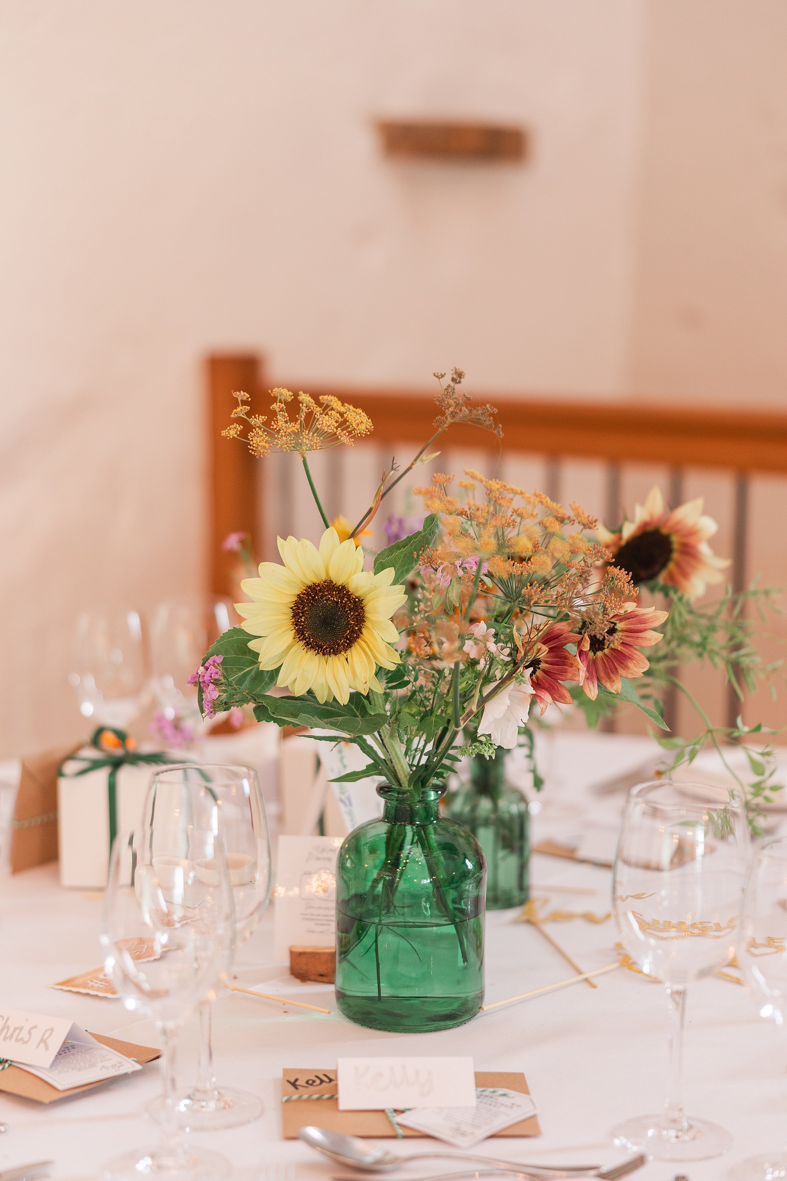 A close up of a vase with a sunflower sitting on a table with a white cloth at a wedding breakfast table