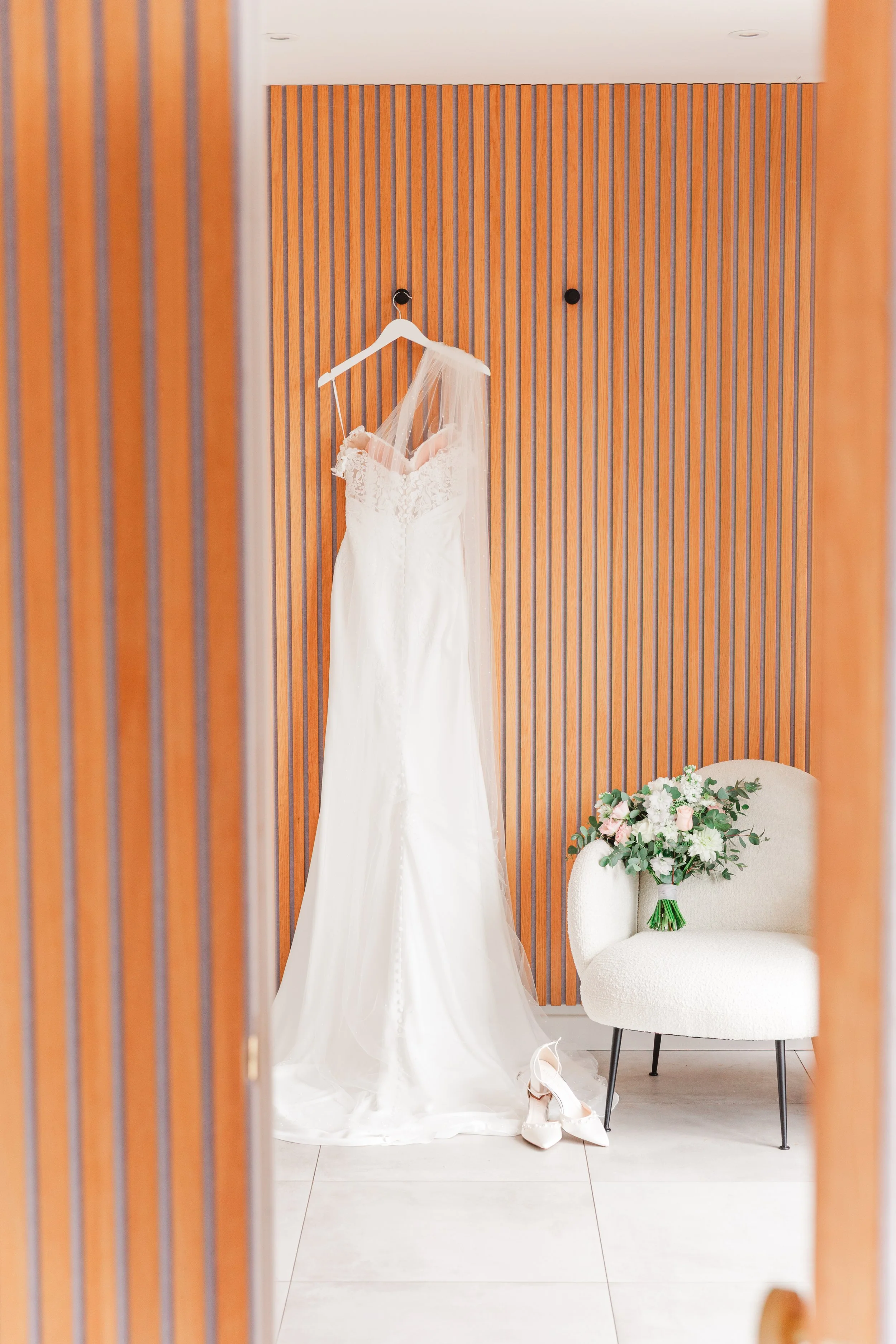 An elegant modern wedding dress hanging in a wooden bridal suite with pretty pastel flowers on a cream chair next to it