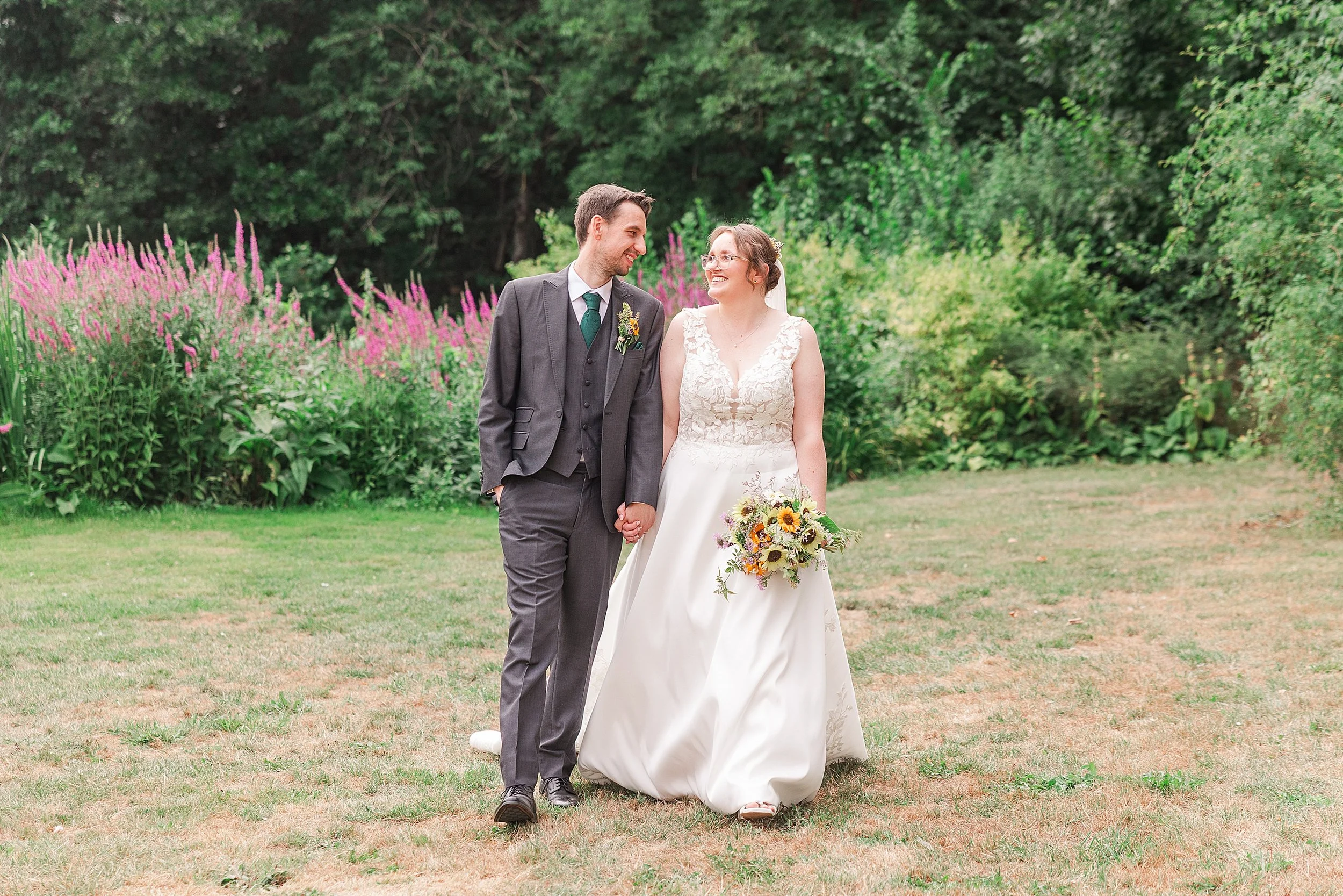 A bride with brown hair and glasses walking hand in hand with her groom with dark hair and a grey suit, they are smiling at eachohter in an English country garden in summer full of lavendar