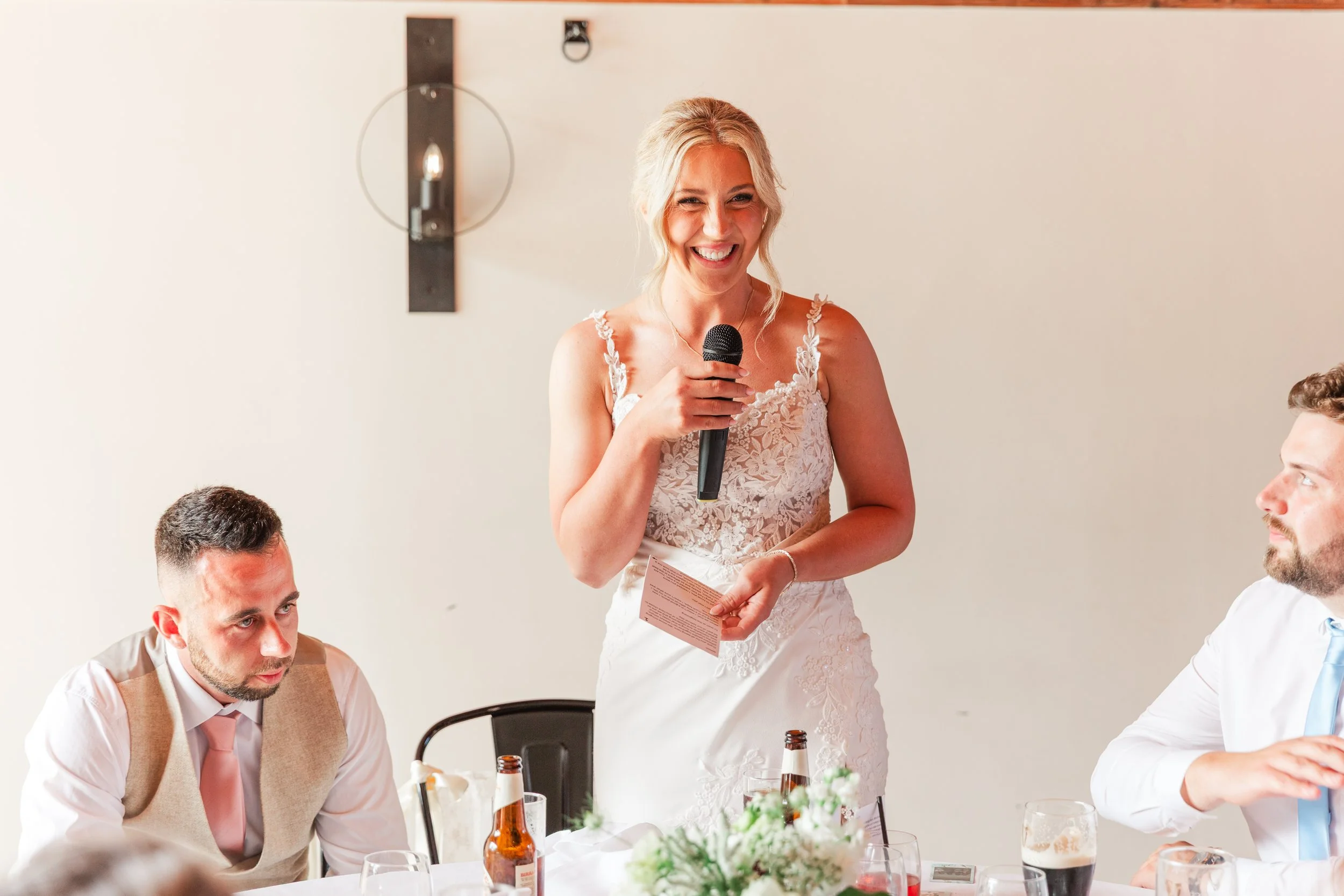 A bride in a modern fitted lace dress laughing giving her speech during a wedding breakfast at Upton Barn