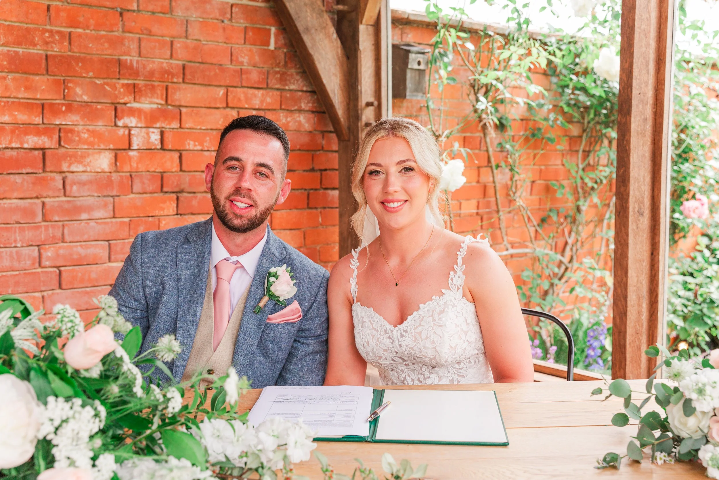A bride and groom smiling at the camera whilst signing their registar