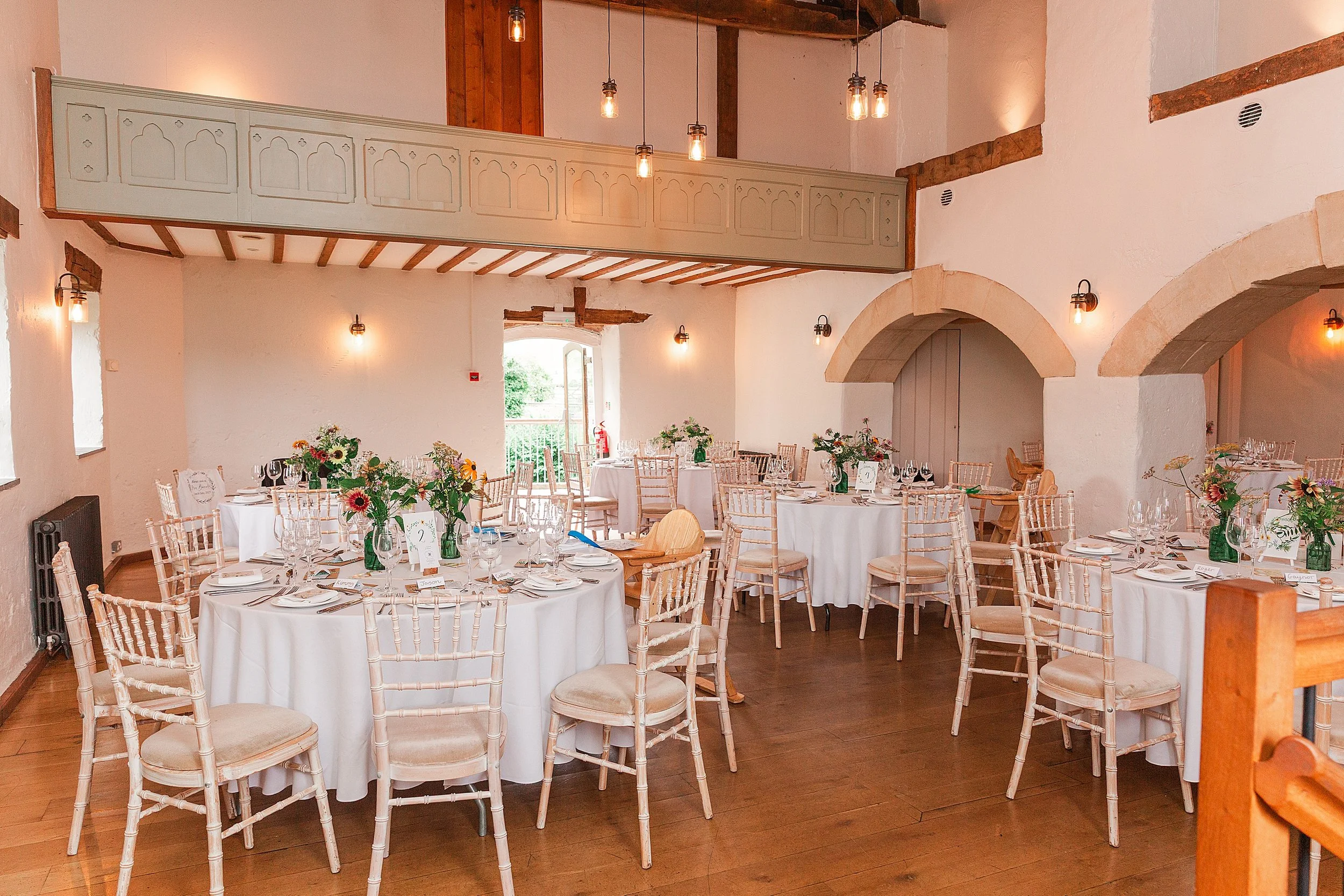 A wedding breakfast room in a barn venue with white walls and wooden beams, there are vases full of summer flowers on the round tables and a wooden floor