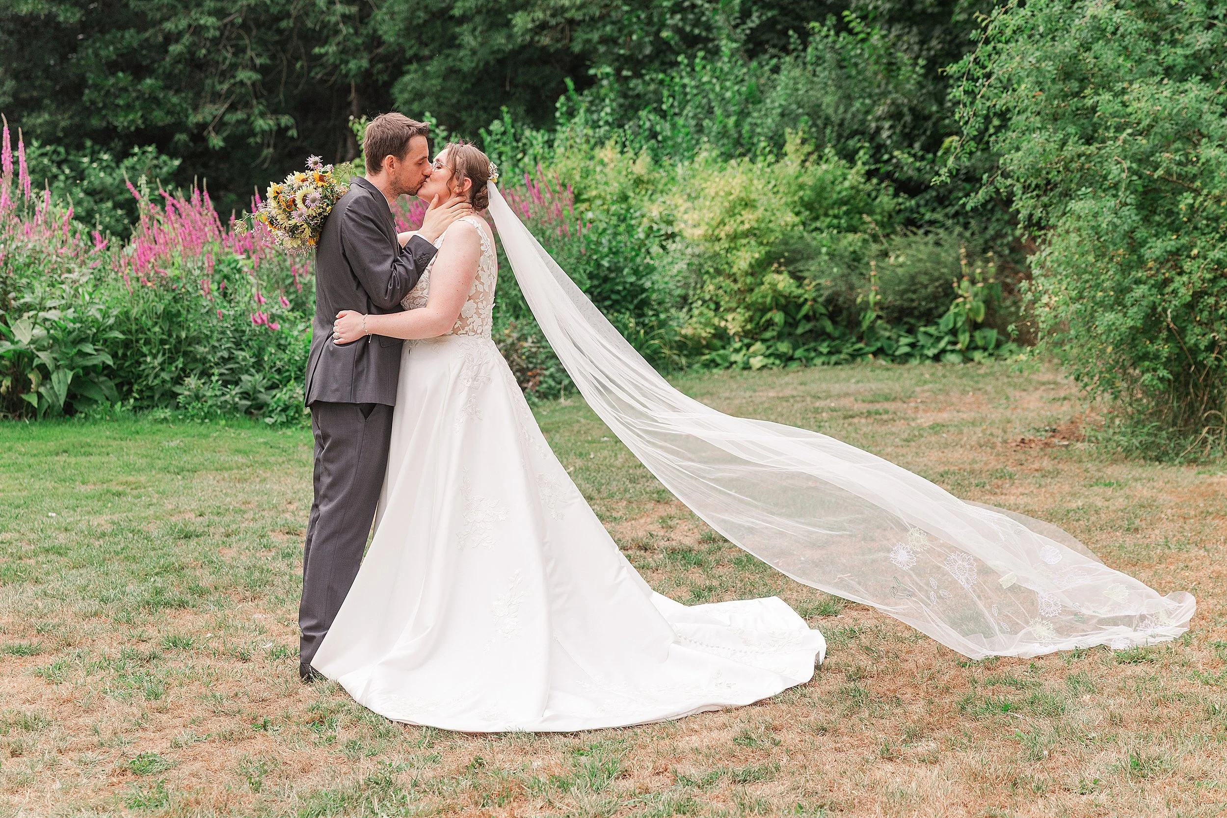 A bride with brown hair kissing her groom with dark hair and a grey suit, the veil os flying high in an English country garden full of lavender