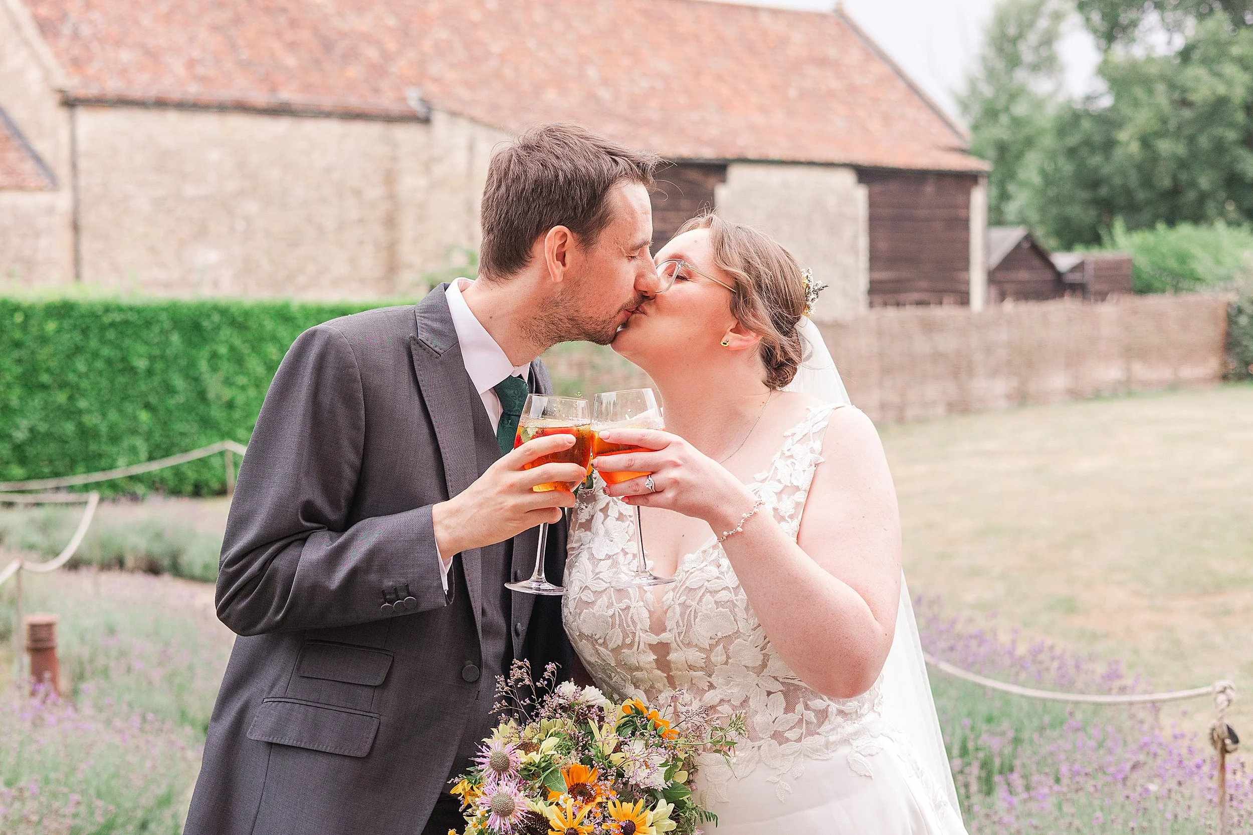 A bride with brown hair and glasses kissing her groom in grey suit whilst cheering a glass of bubbly together