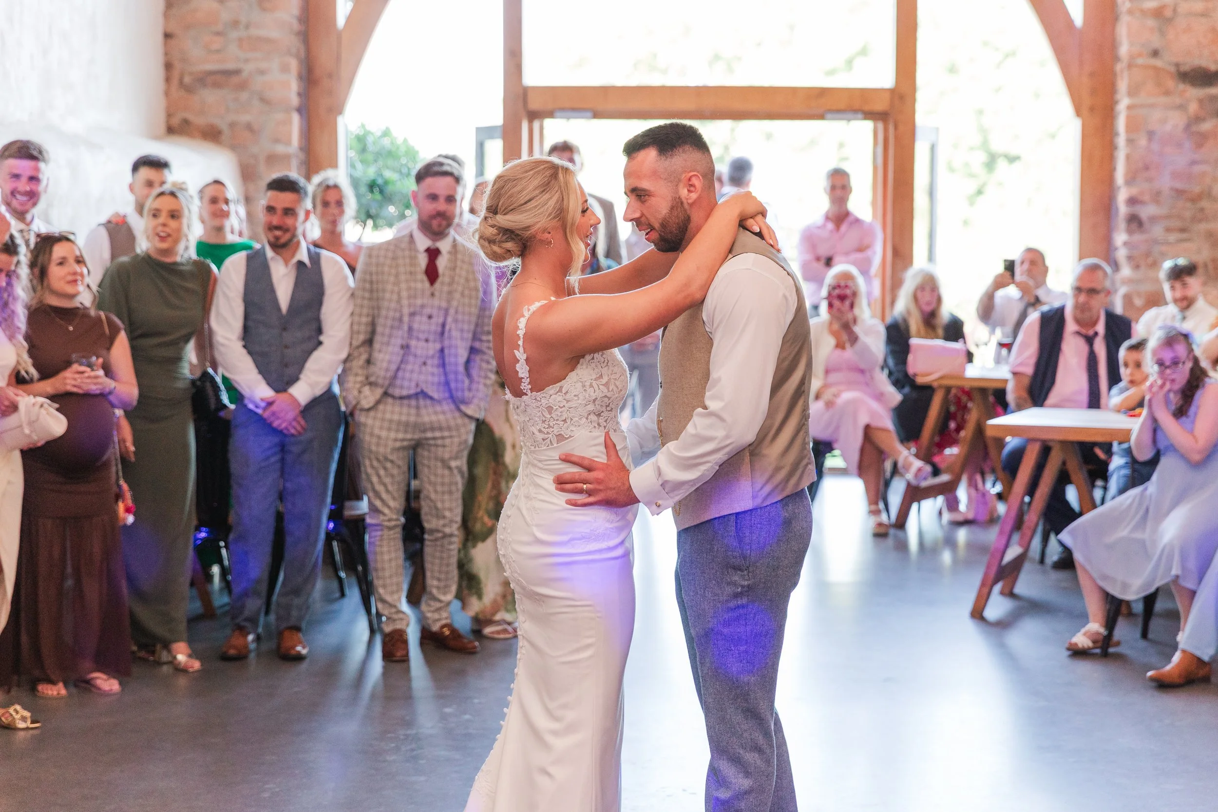 A bride with a fitted lace dress dancing with her groom in a beige waistcoat in a Devon barn with all their guests watching and smiling