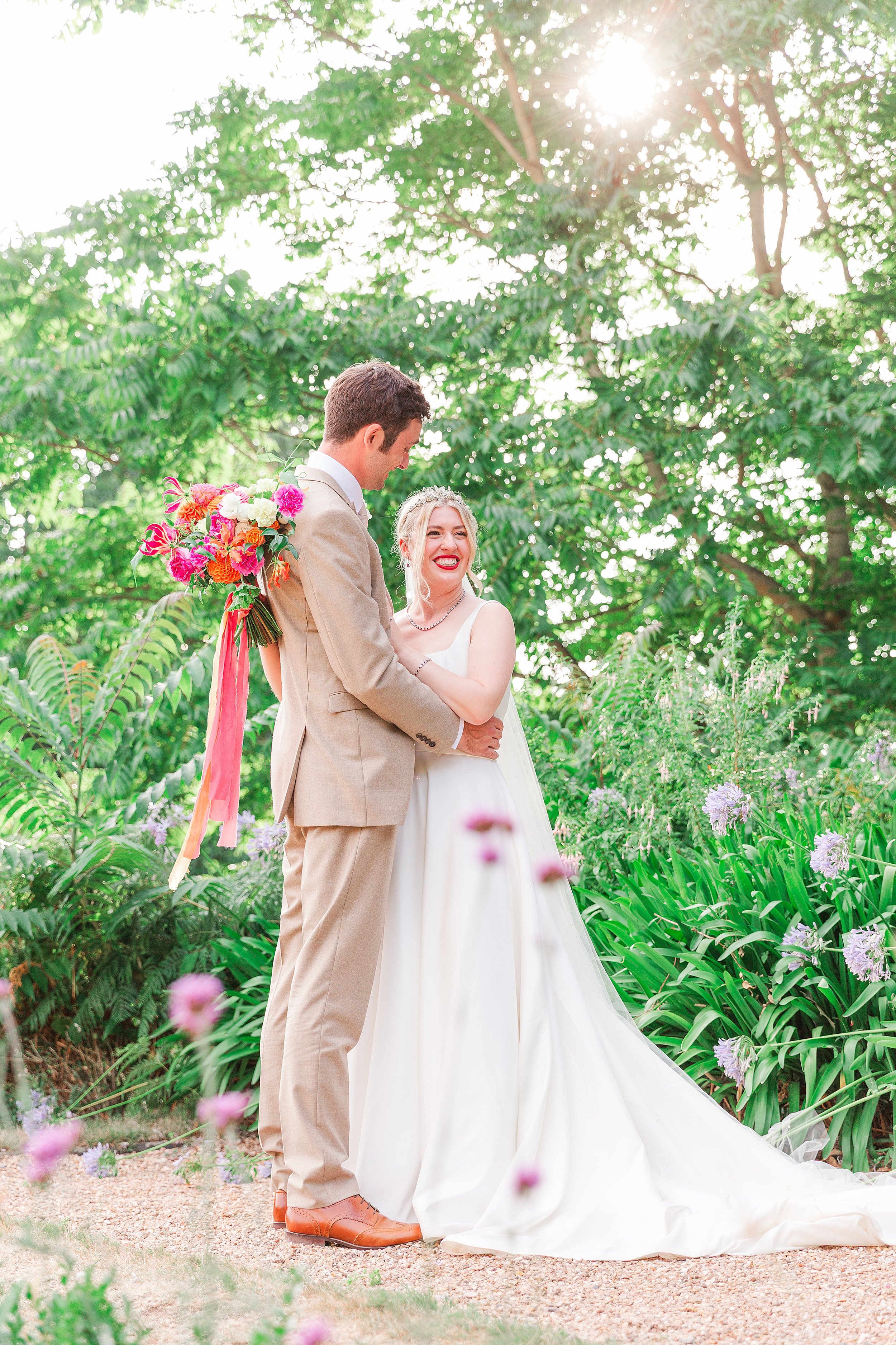 A groom dressed in a stone suit looking at his bride whilst standing among pretty flowers in golden hour