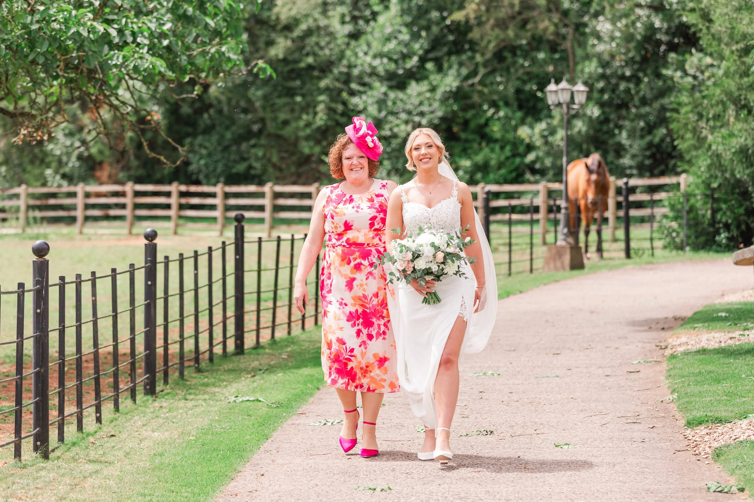 A bride in a modern lace wedding dress walking to her ceremony with her mum in a pink and white dress with a field in the background with a horse