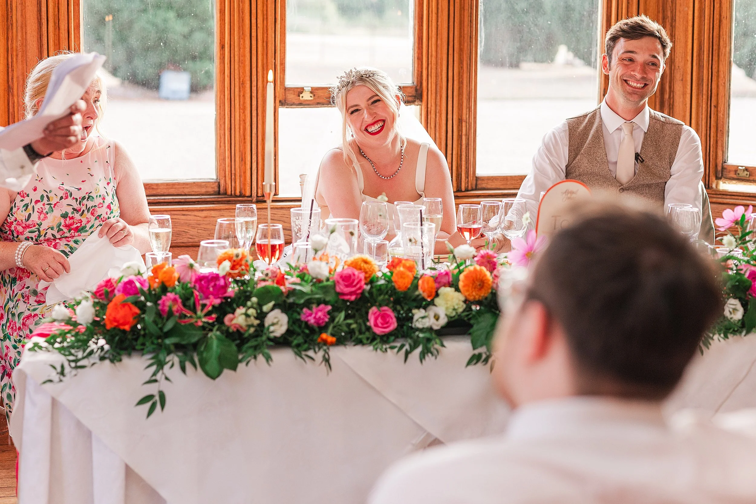 A bride smiling sitting at her wedding breakfast with her groom and mum sat next to her