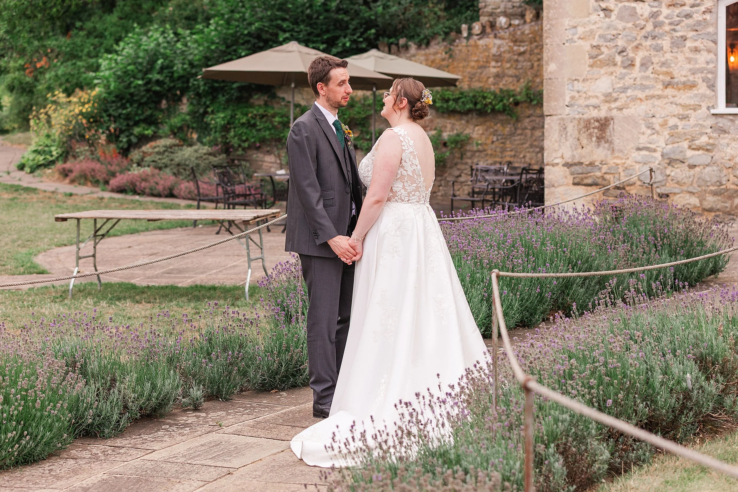 A groom with dark hair and a grey suit holding his brides hands whilst talking in a lavender filled garden