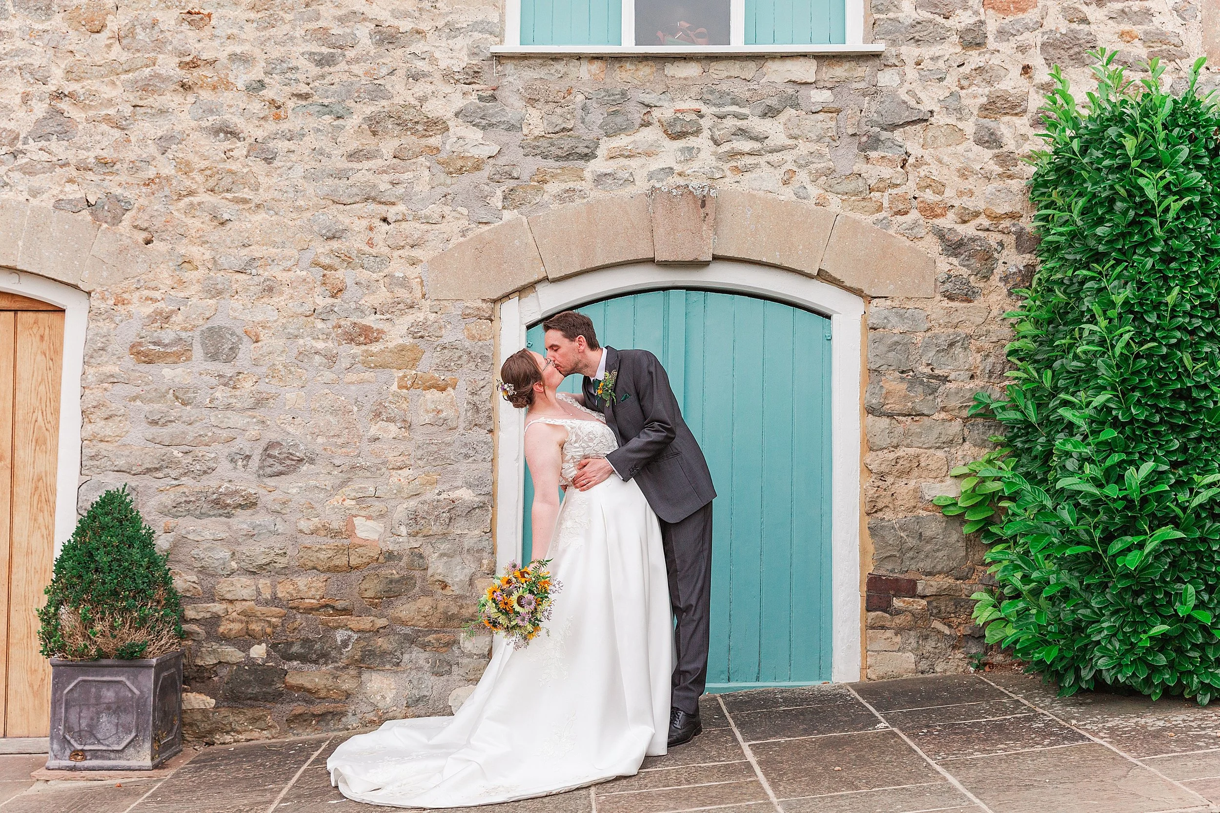 A groom with dark hair and a grey suit dipping and kissing his bride in front of a blue door