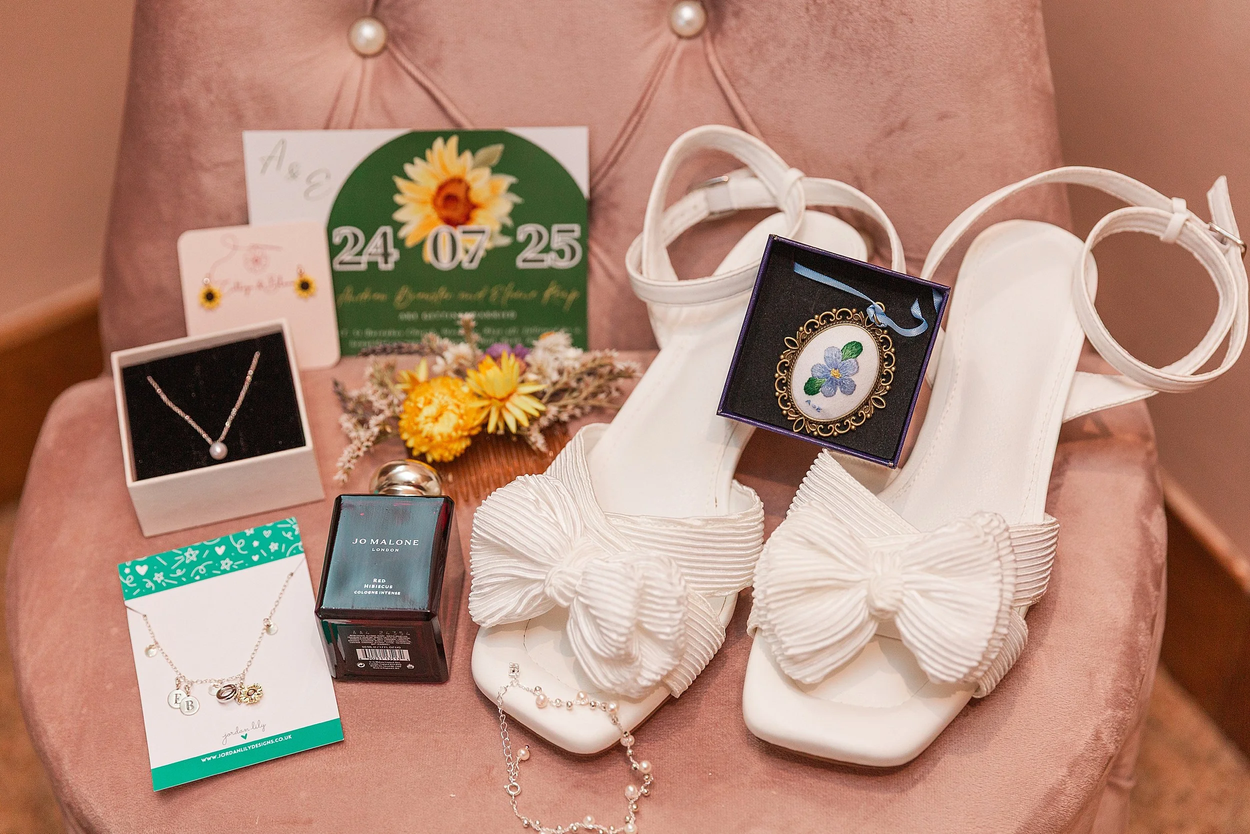 A brides details laid out on a mink coloured chair, there are white shoes, a flower brooch, earrings, necklace and a dried flower hair comb