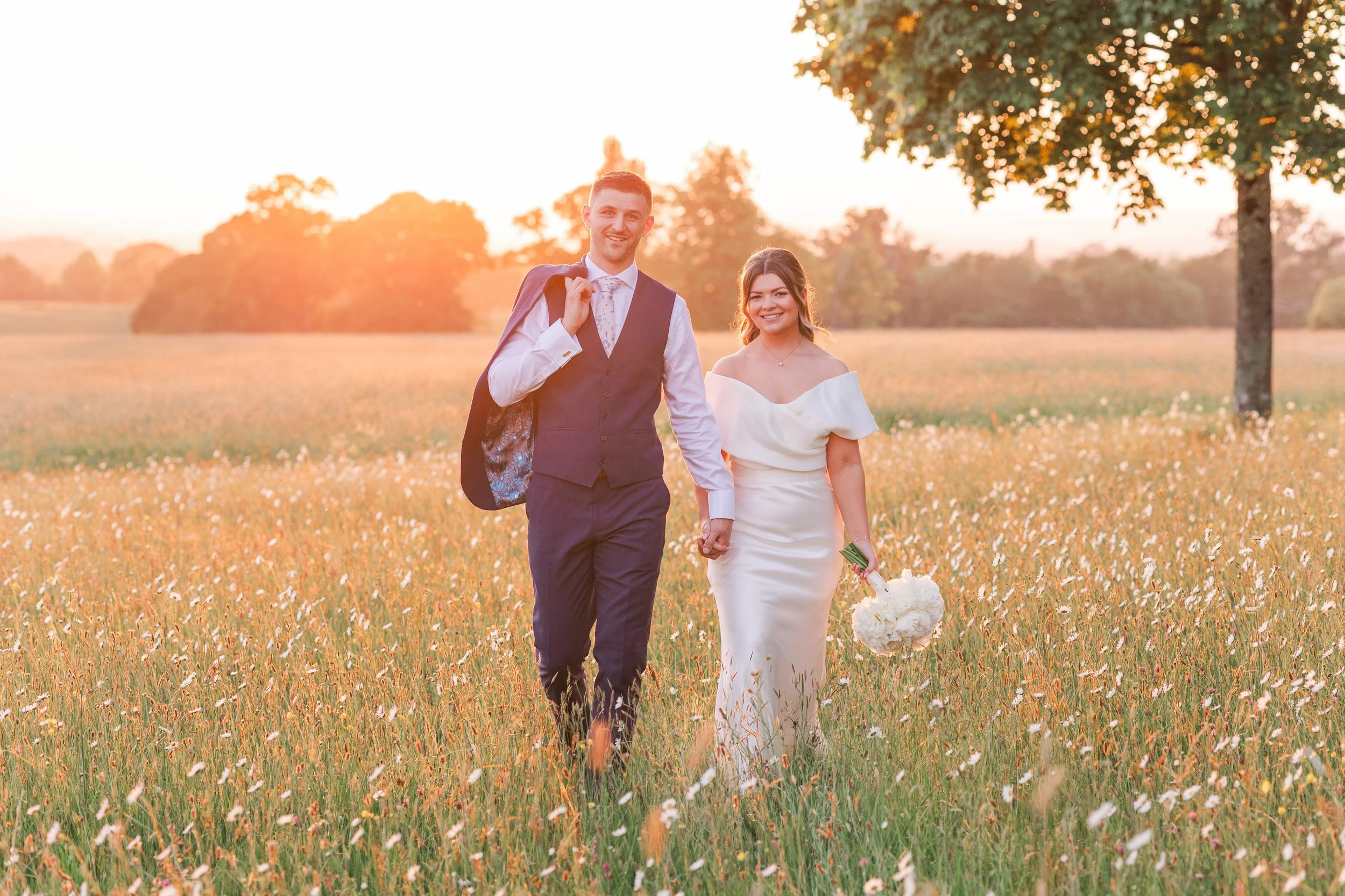A newlywed couple walking hand in hand through a field of wildflowers at sunset. The groom is wearing a navy vest and suit with a white shirt, carrying a jacket over his shoulder. The bride is in a white off-the-shoulder dress, holding a bouquet of white flowers. A large tree is on the right side, and the scene is illuminated by warm, golden sunlight.