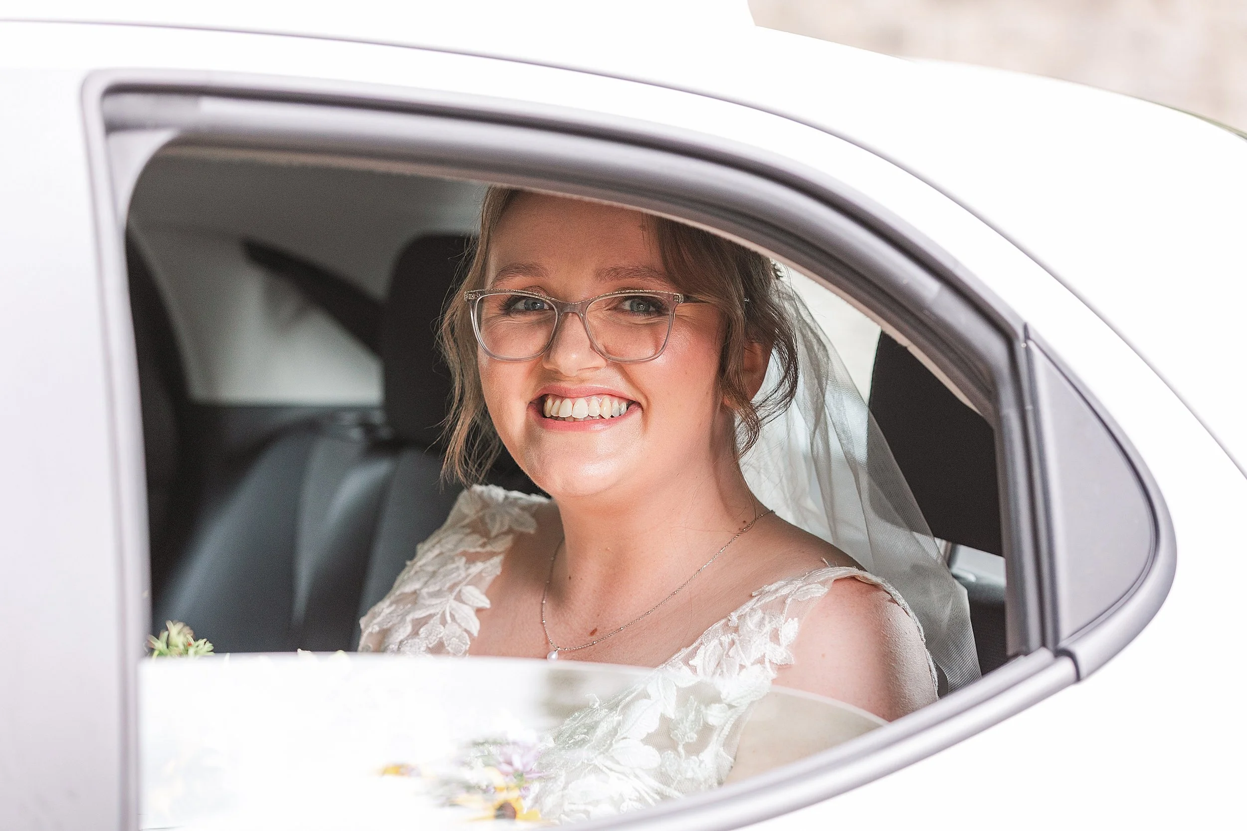 A bride wearing glassed looking out the window of a white car smiling at the camera