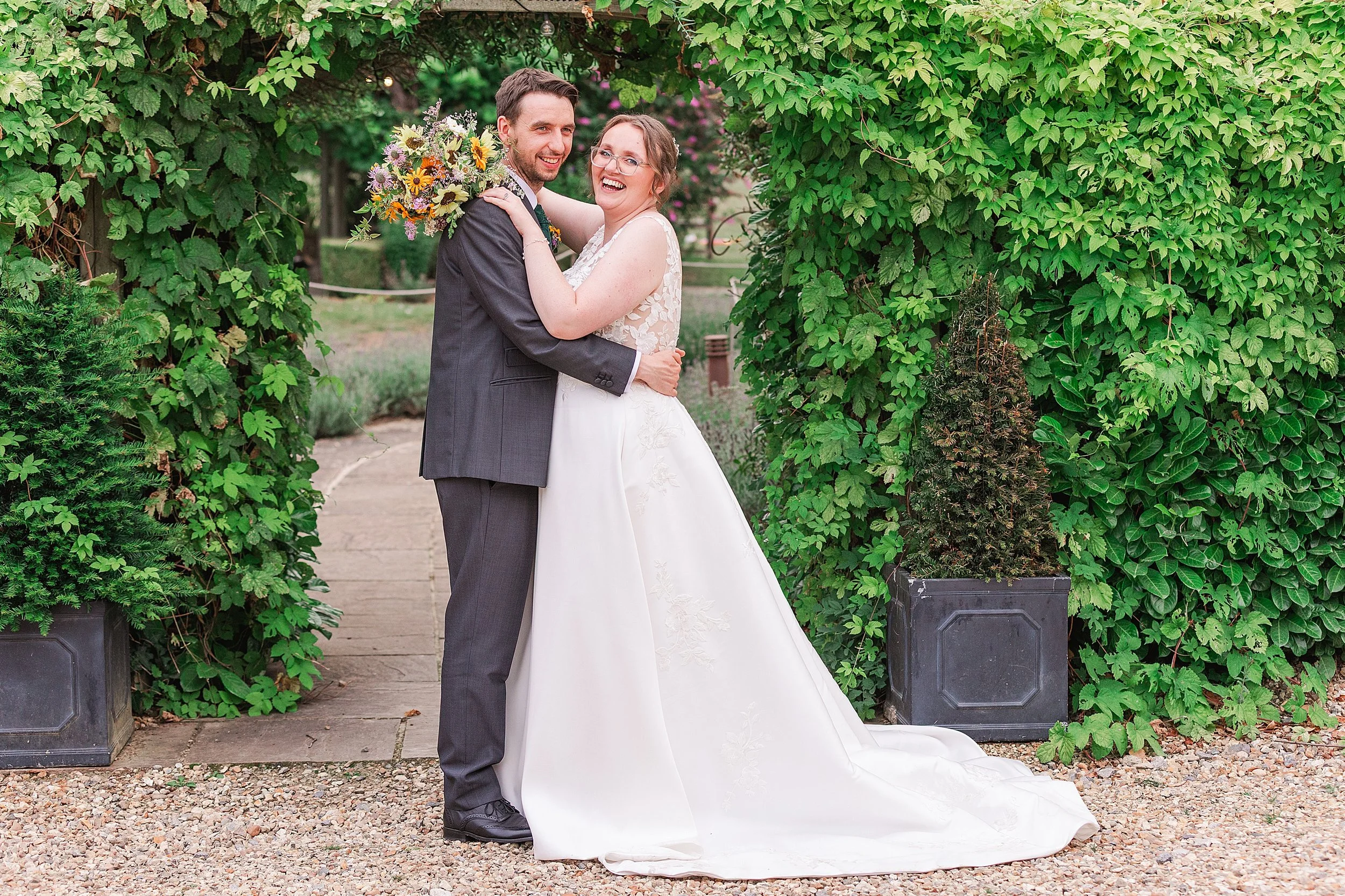 A bride with brown hair and glasses and her groom with dark hair and a grey suit laughing at the camera standing under a foliage filled archway