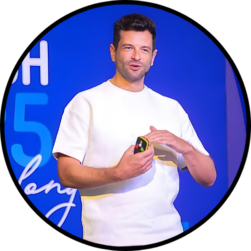 Image of Roy Gluckman on stage delivering a keynote speech. Standing behind a vibrant blue background, holding a clicker