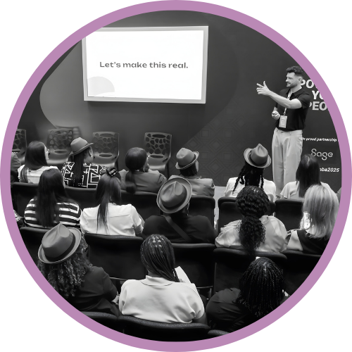 Roy facilitating a training session. Roy is standing at the front and the audience is looking at him. At the back is a creen that says 'Let's Make this real'.