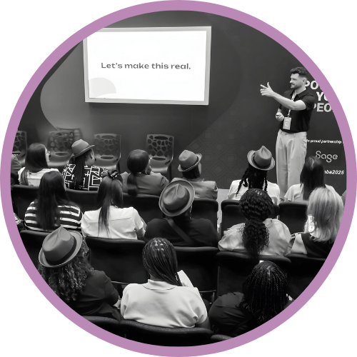 Roy facilitating a training session. Roy is standing at the front and the audience is looking at him. At the back is a screen that says 'Let's Make this real'.