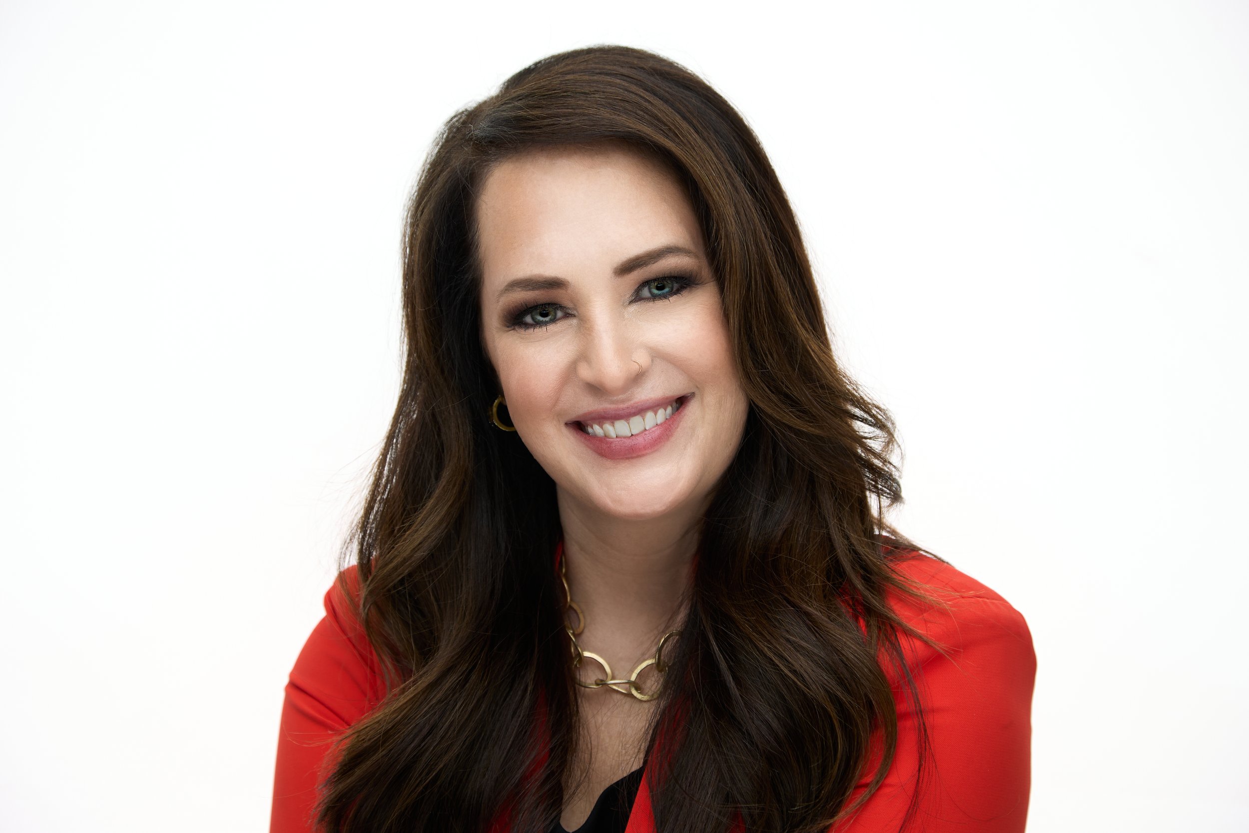 A woman with long dark brown hair smiling, wearing a red blazer and gold jewelry, against a plain white background.