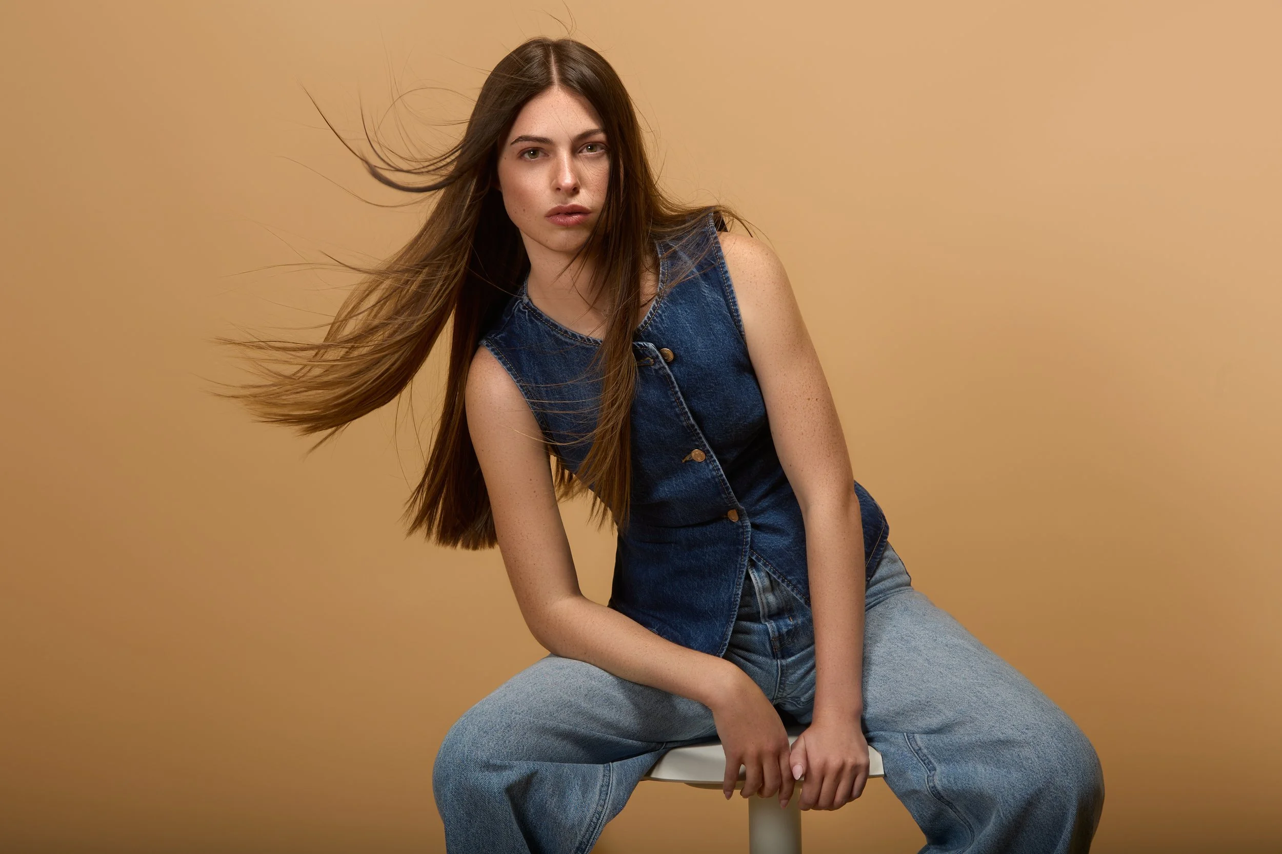 Young woman with long flowing hair wearing a sleeveless denim vest and baggy jeans, sitting on a stool against a beige background.