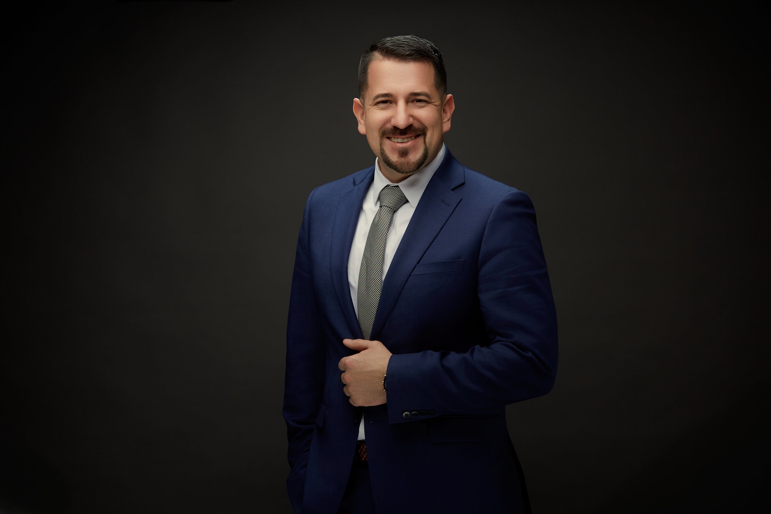 A man in a blue suit, white shirt, and gray tie posing against a black background, smiling with short dark hair and a trimmed beard.