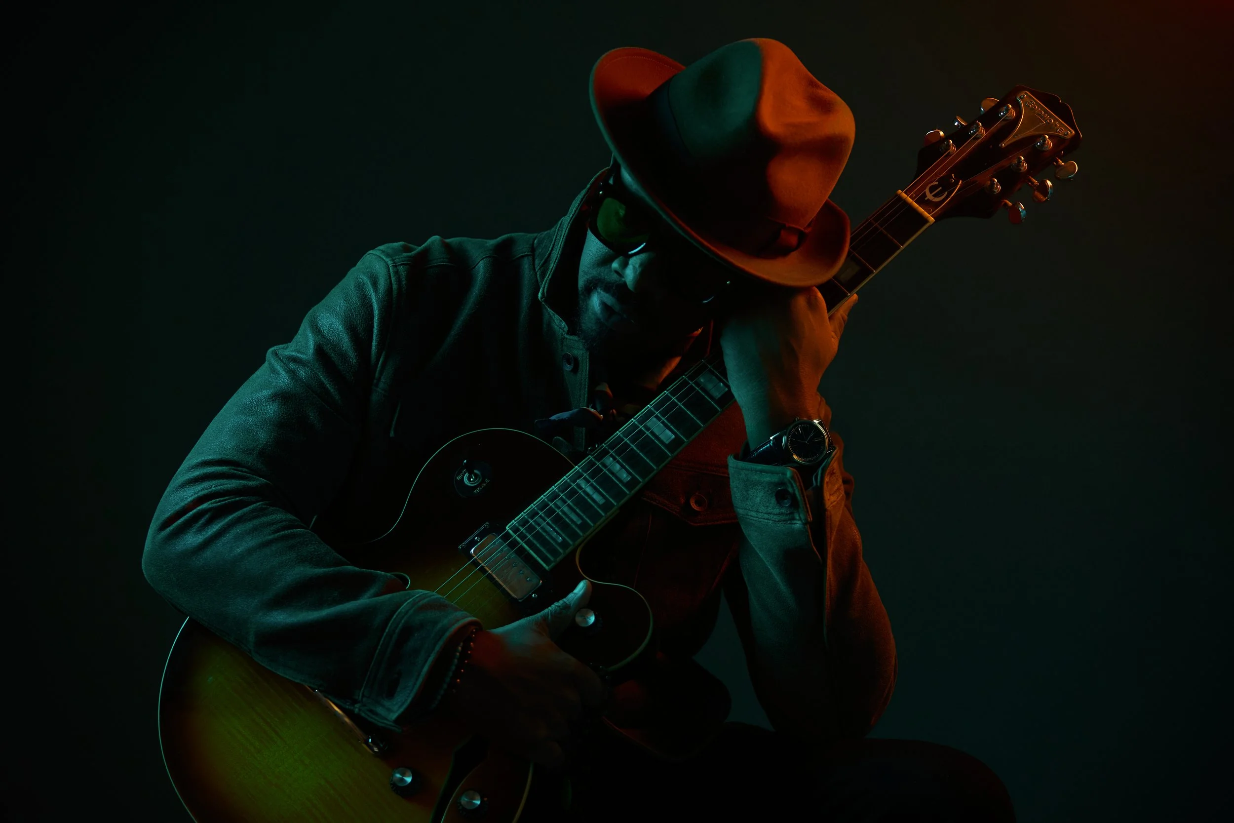 Musician wearing a brown fedora hat and dark sunglasses, holding an electric guitar in a dimly lit setting.