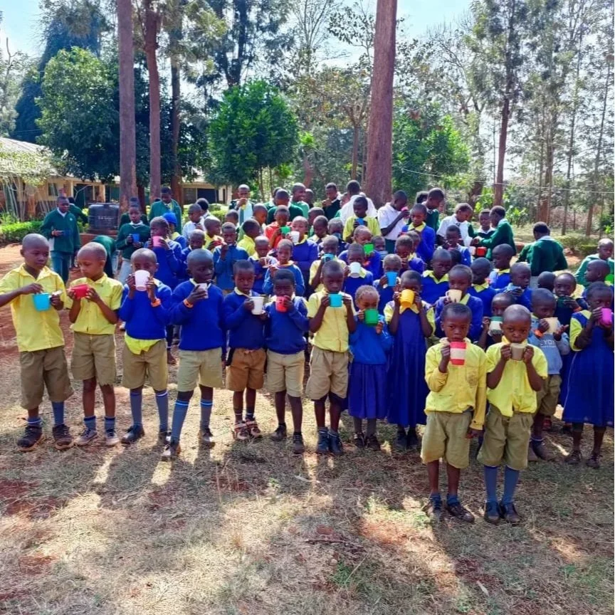 A large group of Kenyan school children holding mugs of porridge, standing in front of trees.