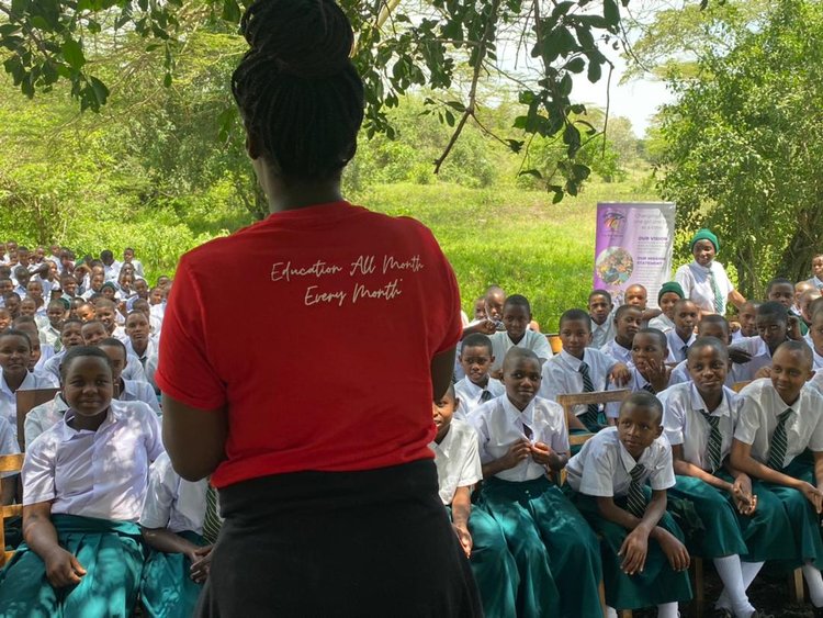 The back of a lady in a red t-shirt with an 'Education All Month, Every Month' slogan addressing a group of school children