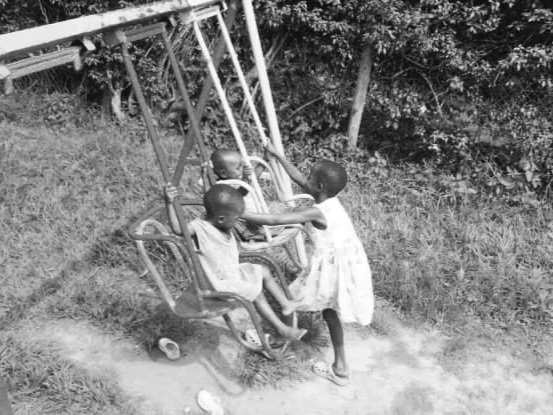 A black & white photo of 3 Ugandan children playing on rudimentary swings.