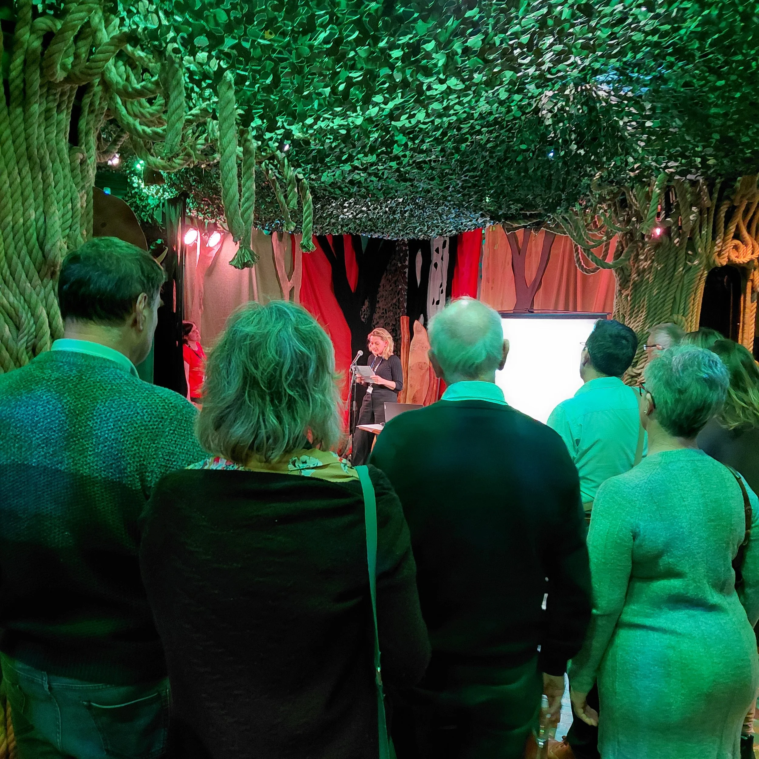 A blonde-haired lady speaking into a microphone, as a crowd gathers round to listen to her.